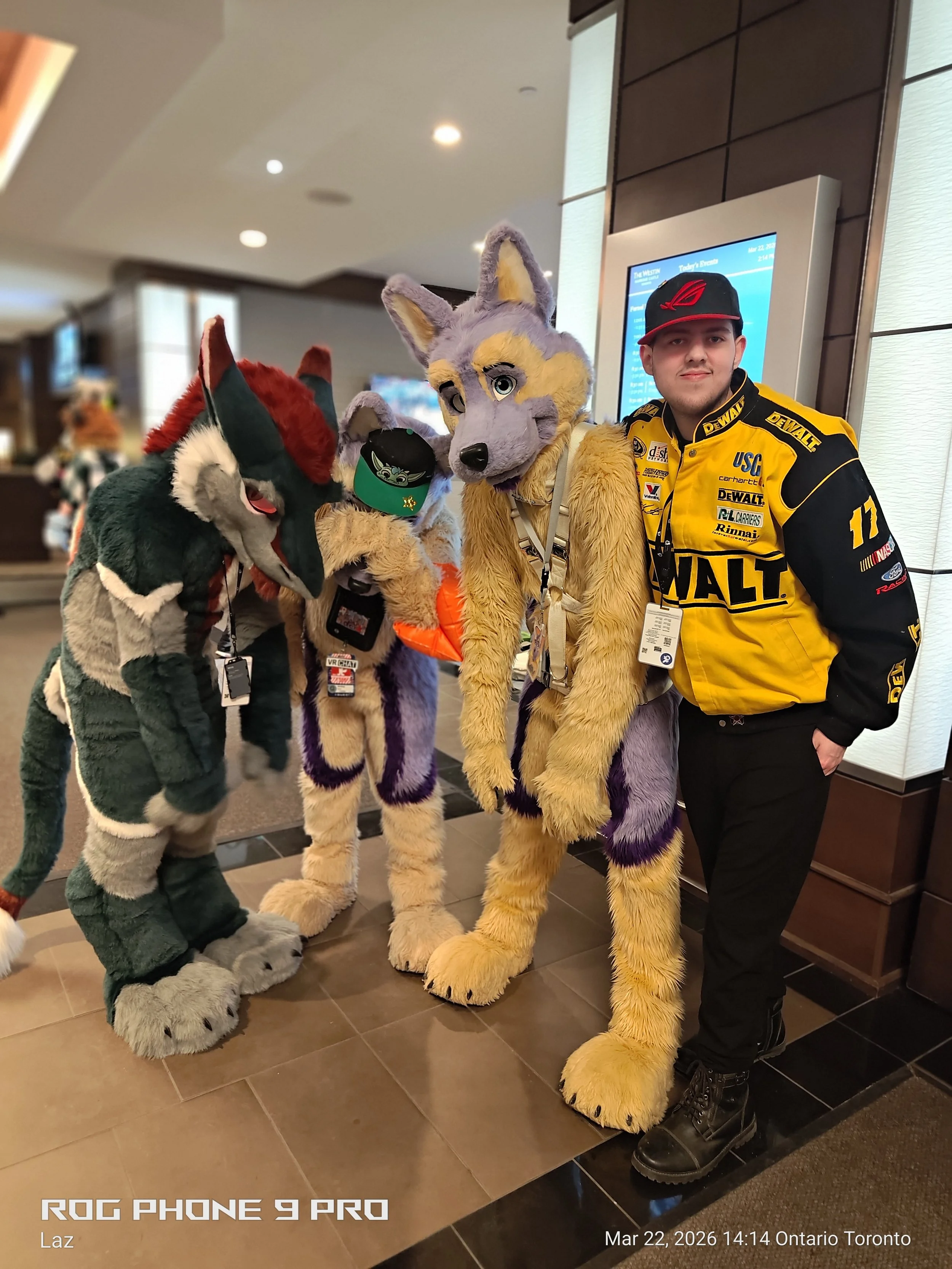 A man in a yellow racing jacket and black pants stands pridefully next to three people dressed in colorful animal costumes, including a wolf, a fox, and a chipmunk, in what appears to be a hotel lobby or conference center.