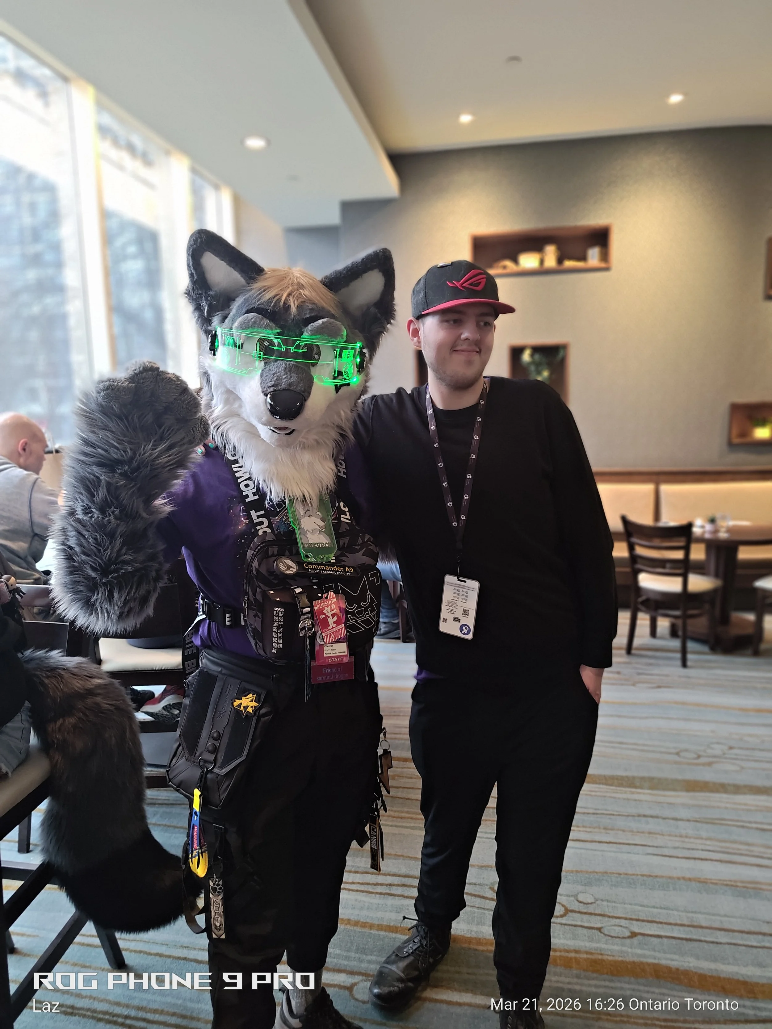 A person in a wolf costume with green visor glasses standing next to a man wearing a black shirt and a cap, inside a well-lit room with modern decor.