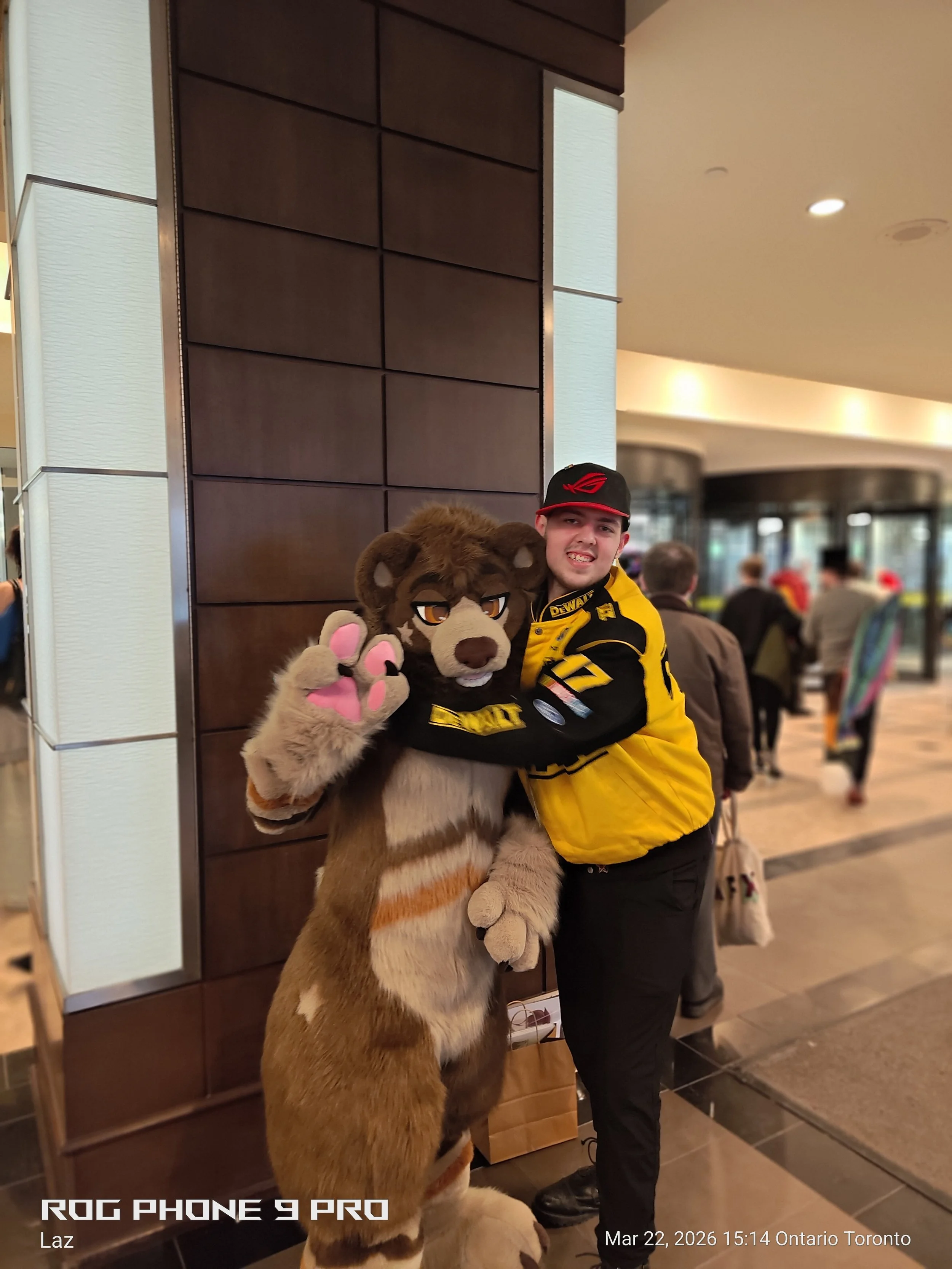 A person in a yellow and black jacket and a black cap stands next to a person in a lion mascot costume. The mascot is giving a paw with pink pads. They are in a busy indoor public space, possibly a hotel or conference center, with people in the backg