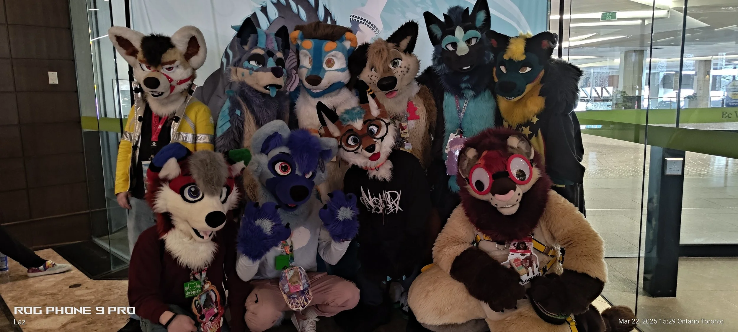 Group of people in colorful animal fursuits posing for a photo in a lobby with glass walls.