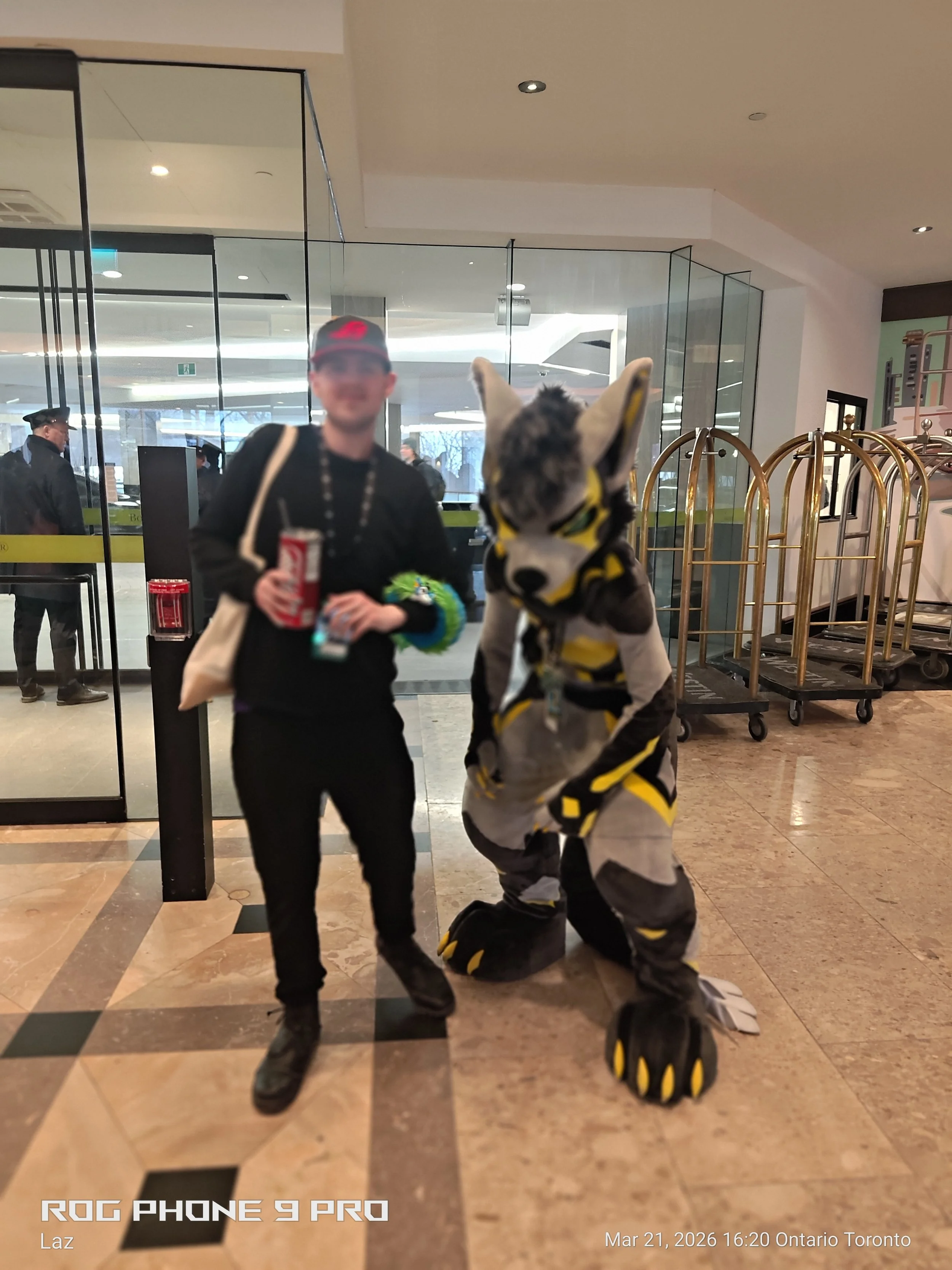 A person dressed in a wolf costume standing on the floor of a shopping mall, holding a stuffed animal and bowing to a person holding a drink.