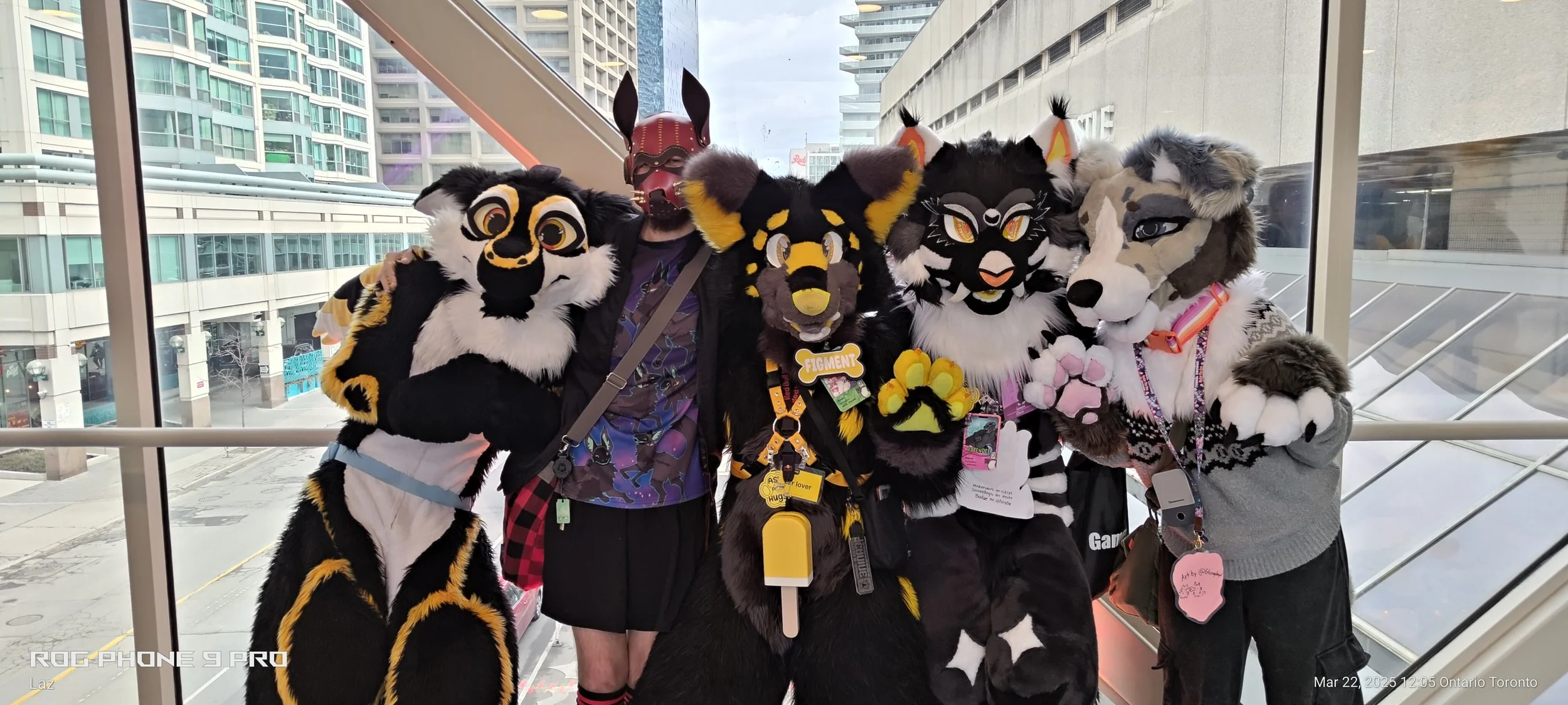 Five people in colorful, detailed furry costumes stand inside a glass-walled building, with a cityscape featuring tall buildings in the background.