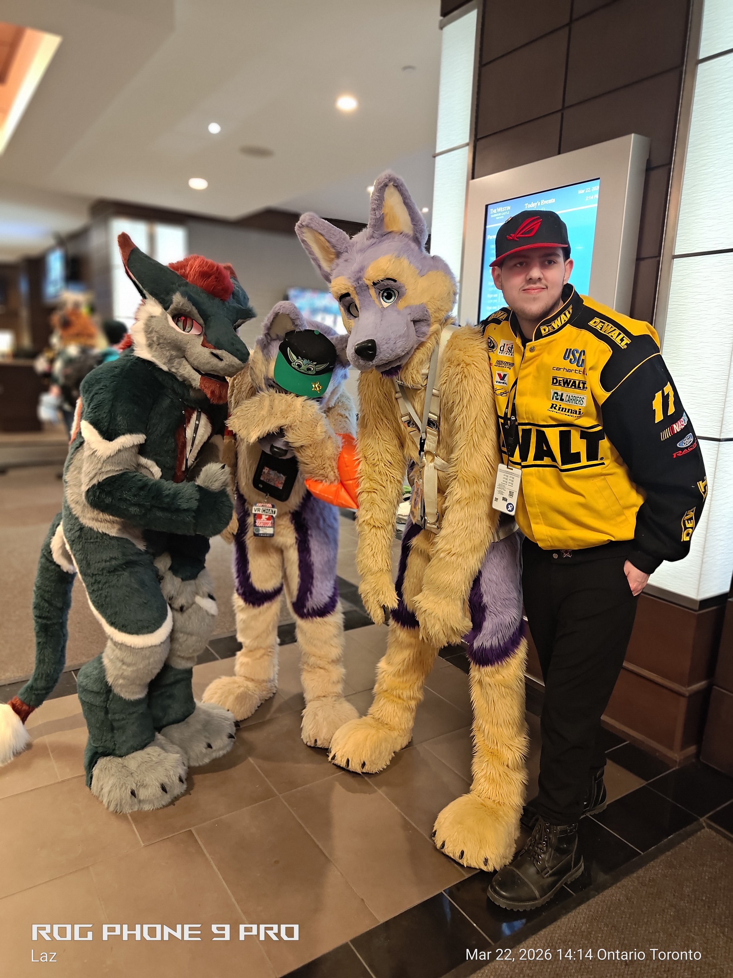 A man in a yellow racing jacket and black pants wearing a black cap stands next to three people in fursuits of anthropomorphic animals, inside a building with a digital screen in the background.