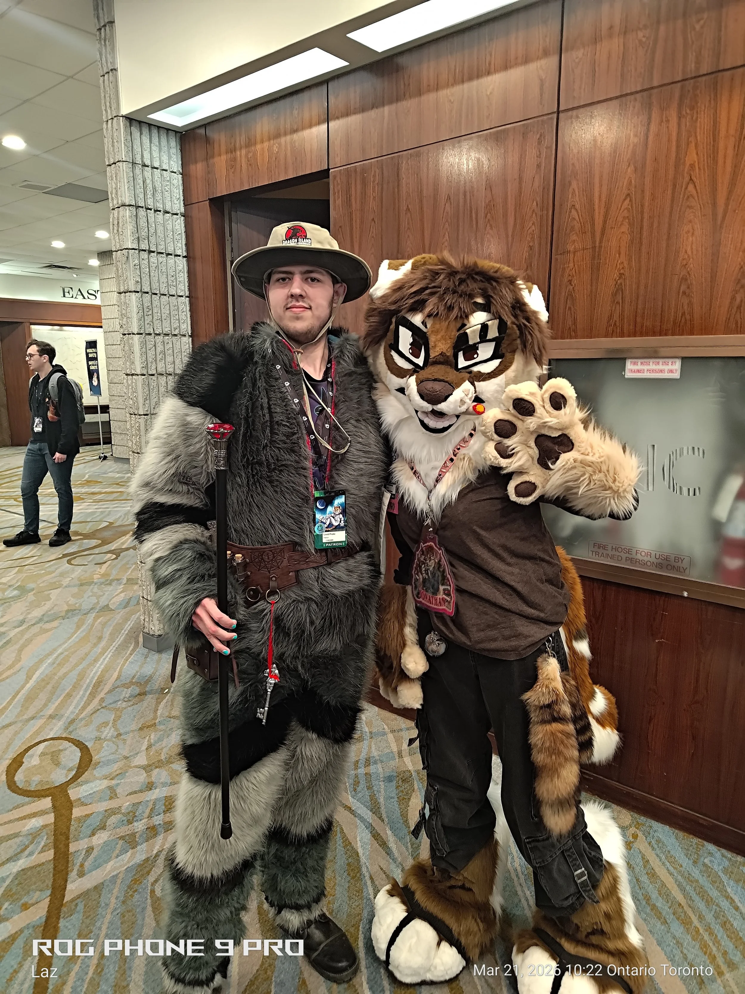 Two people dressed in furry costumes resembling animals pose for a photo at an indoor event. One costume appears as a wolf or some similar animal with gray and black fur, and the other as a feline with brown and white fur. The person in the wolf cost
