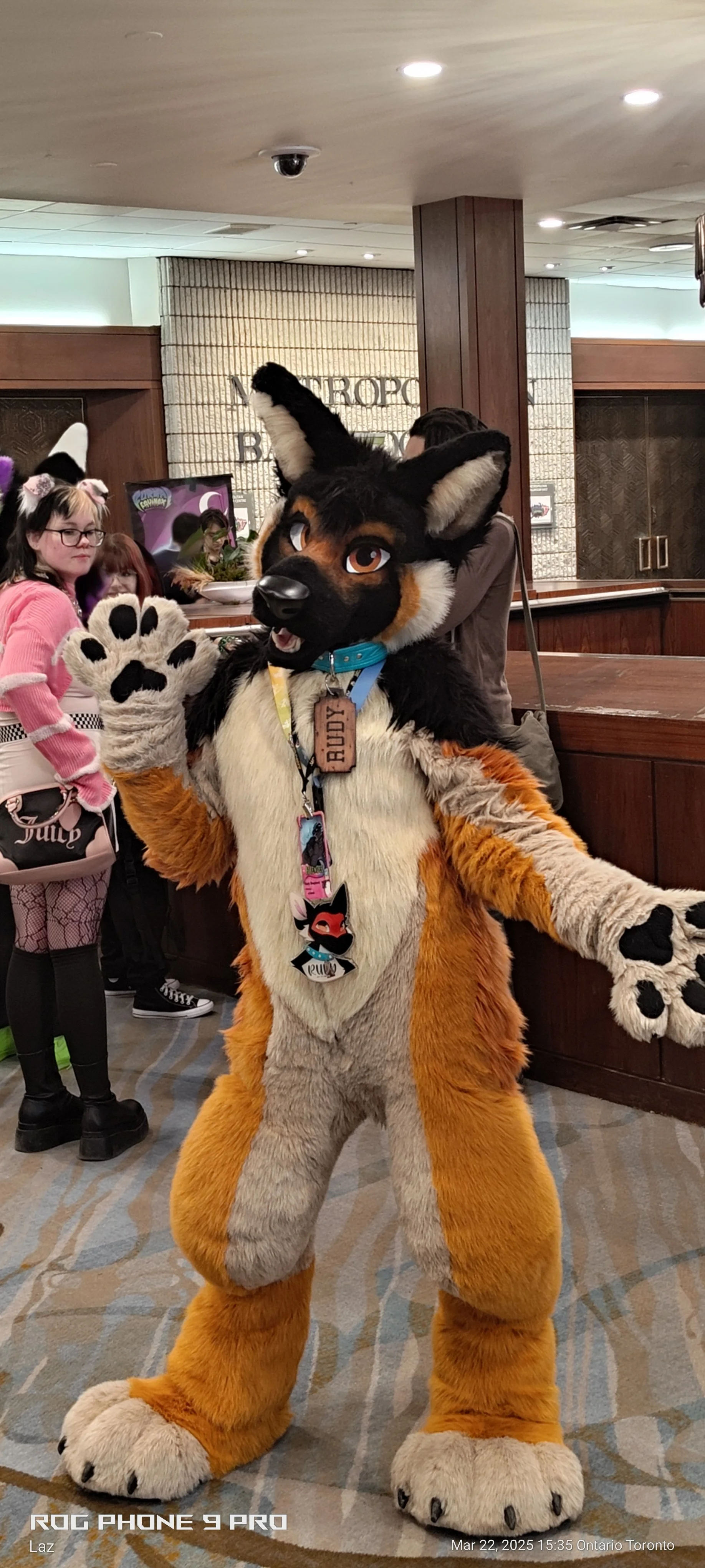 Person in an anthropomorphic fox fursuit waving indoors at a convention or event, with a group of people and a reception desk in the background.