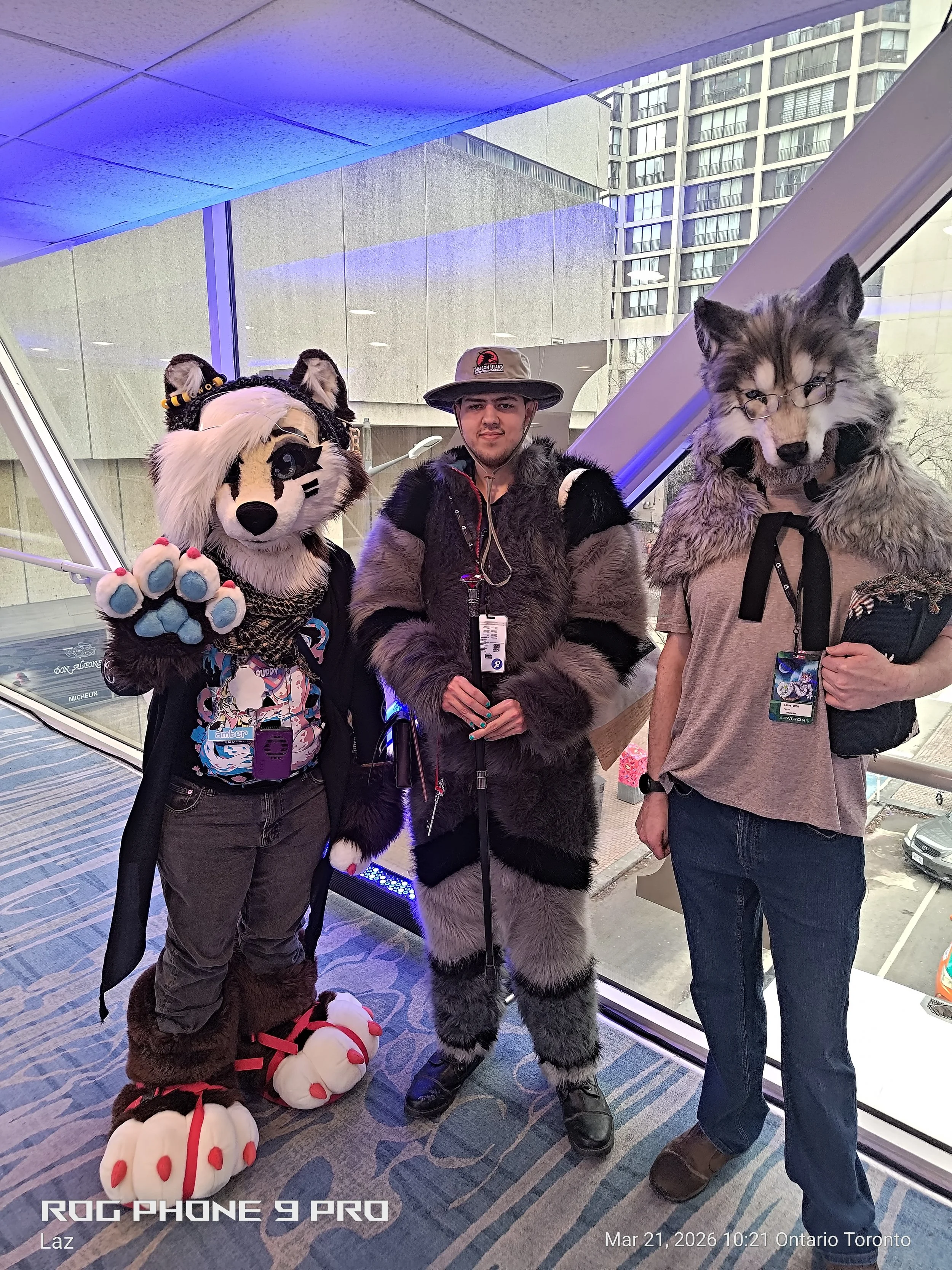 Three individuals dressed in animal costumes posing inside a building, with large windows showing tall buildings outside. One is a panda fursuit, another is a wolf with glasses, and the third is a stylized tiger.