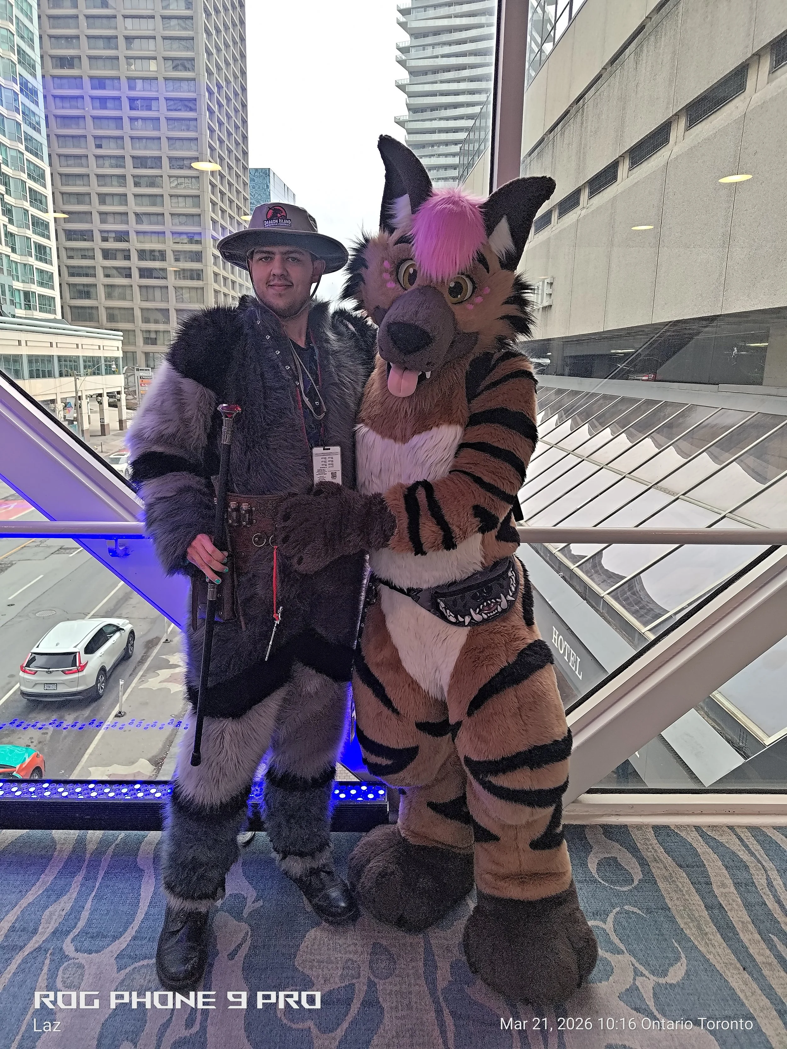 Two people in costume, one dressed as a furry character with pink hair and black stripes, and the other as a large animal character with gray and black fur, standing inside a building near a window with a cityscape view of tall buildings and a parkin