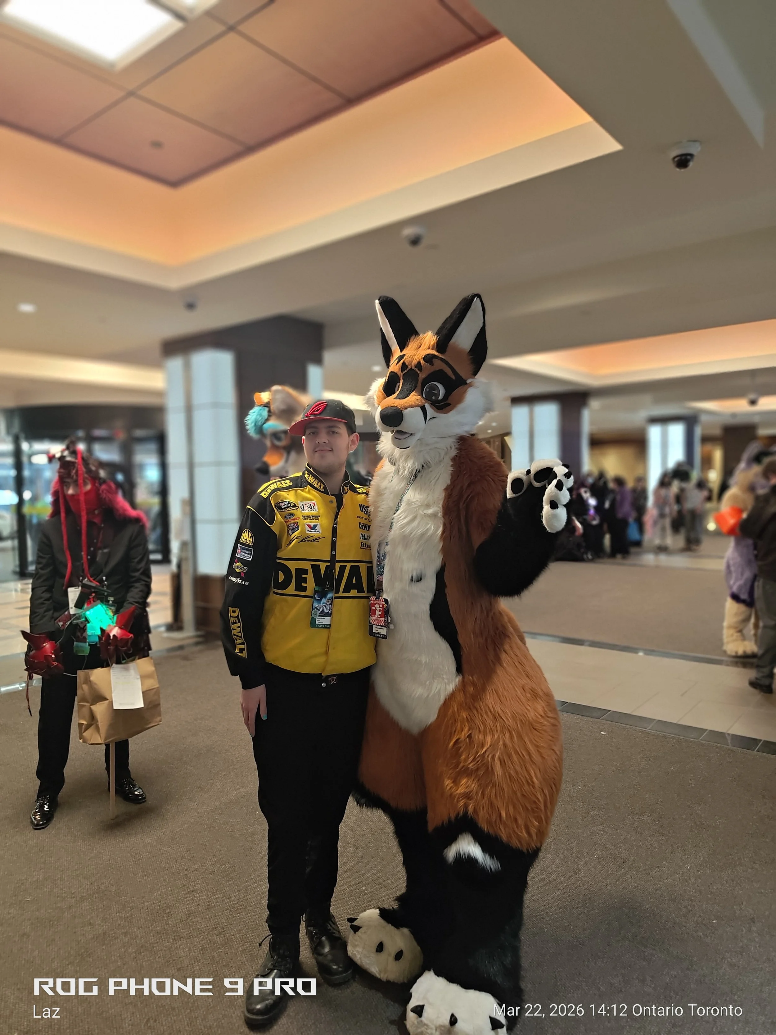 A person wearing a yellow racing jacket standing next to a person in a full-body furry costume of an animated animal, likely a fox or similar creature, in an indoor public space. The person in costume is waving at the camera. There are other people w