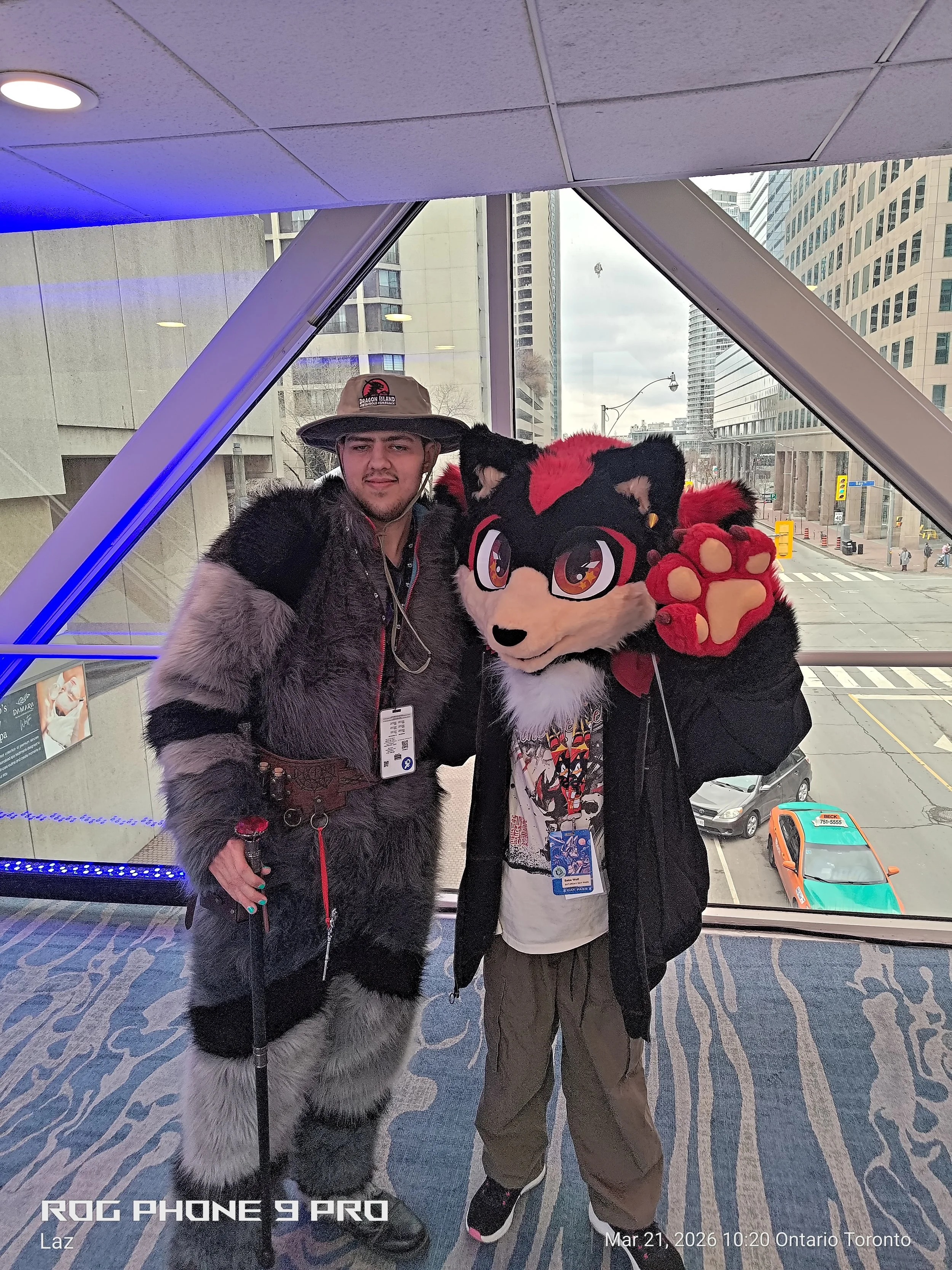 Two people dressed in furry costumes are standing inside a building with a city view outside the window. One is in a wolf costume with gray, black, and white fur, and the other in a red and black costume with a large furry head with glasses and red p