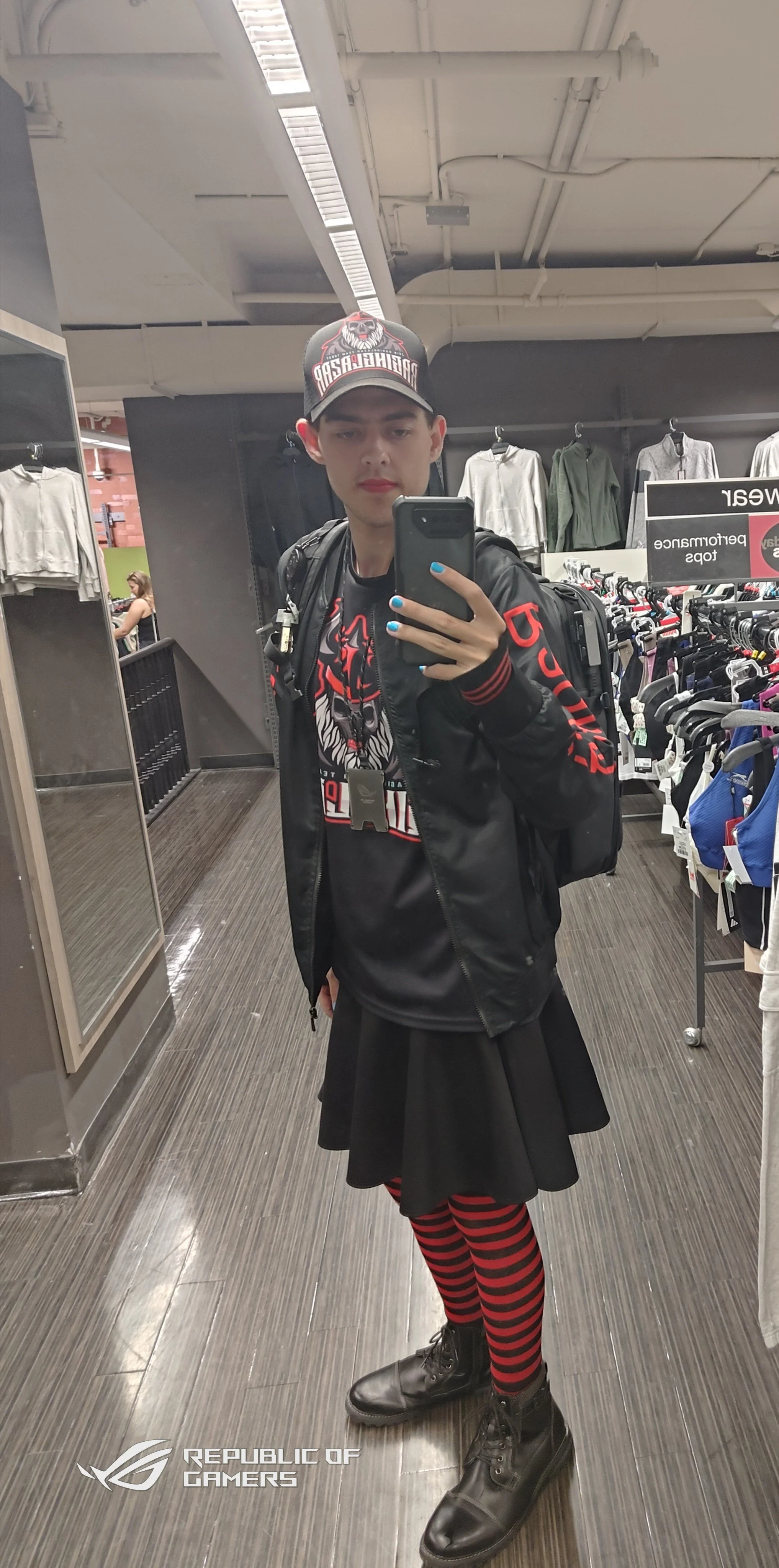 A young person taking a mirror selfie in a clothing store, wearing a black jacket with red lettering, a graphic t-shirt, a black skirt, red and black striped tights, black combat boots, and a black cap with a skull logo. The store has racks of clothi