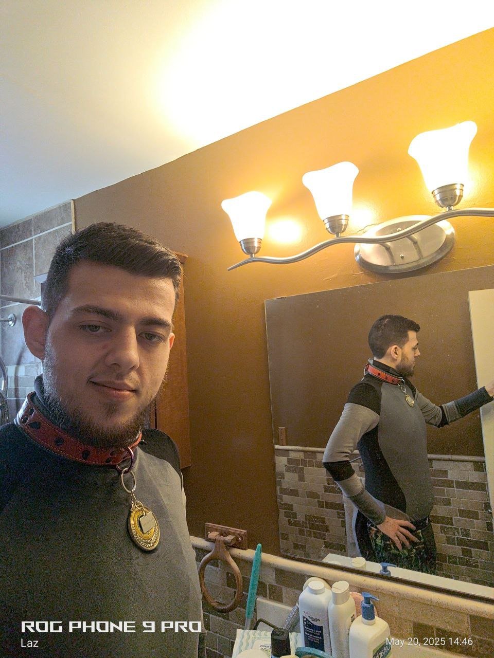 A young man with short brown hair and a beard taking a mirror selfie in a bathroom. He is wearing a gray shirt with black sleeves, a red collar with black holes, and a gold medal necklace. The bathroom has tiled walls, a wooden cabinet, a mirror, a f