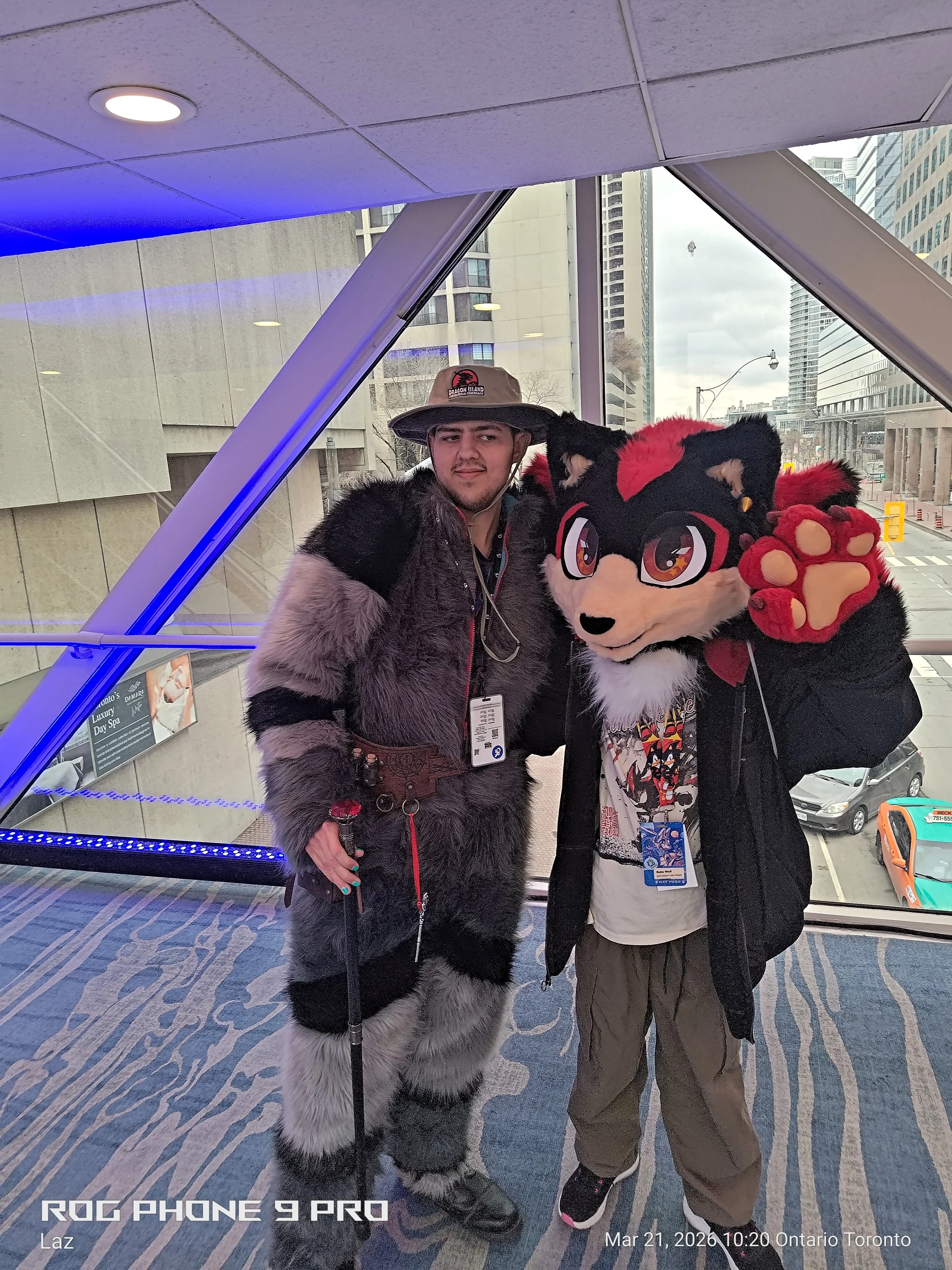 Two individuals are standing together in a modern indoor setting with large windows. One is dressed as a furry mascot with a large black, red, and white costume featuring big animated eyes and paw-like hands. The other is a person wearing a brown hat