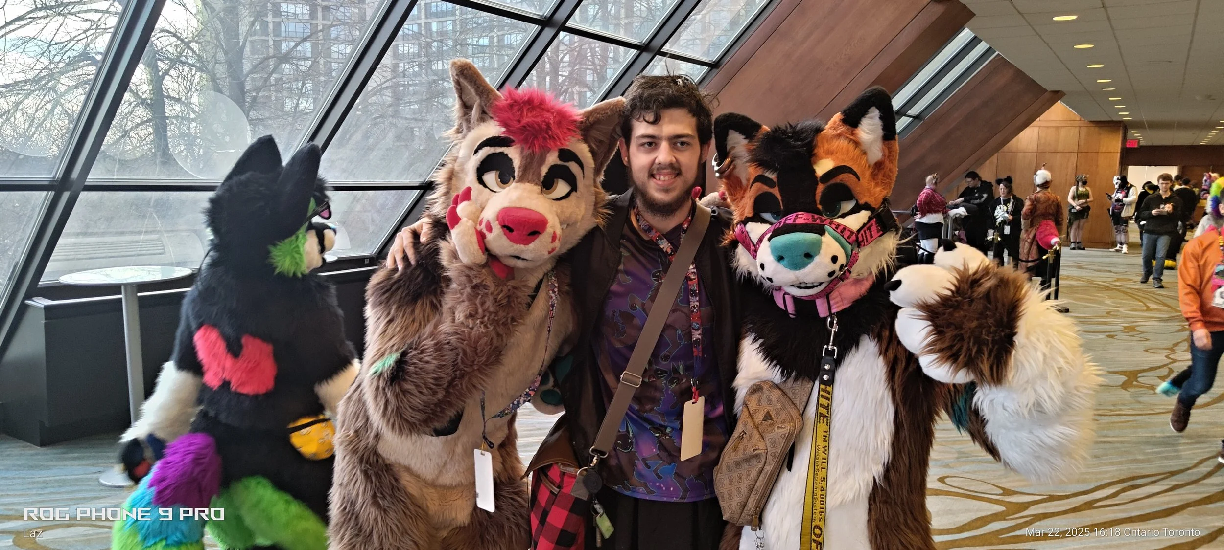 A man posing with three people dressed in colorful furry animal costumes at a convention or event indoors near large windows.