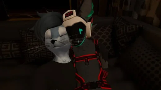 Two cats wearing virtual reality headsets, one with a white face and black markings, the other with a black face, seated on a couch.