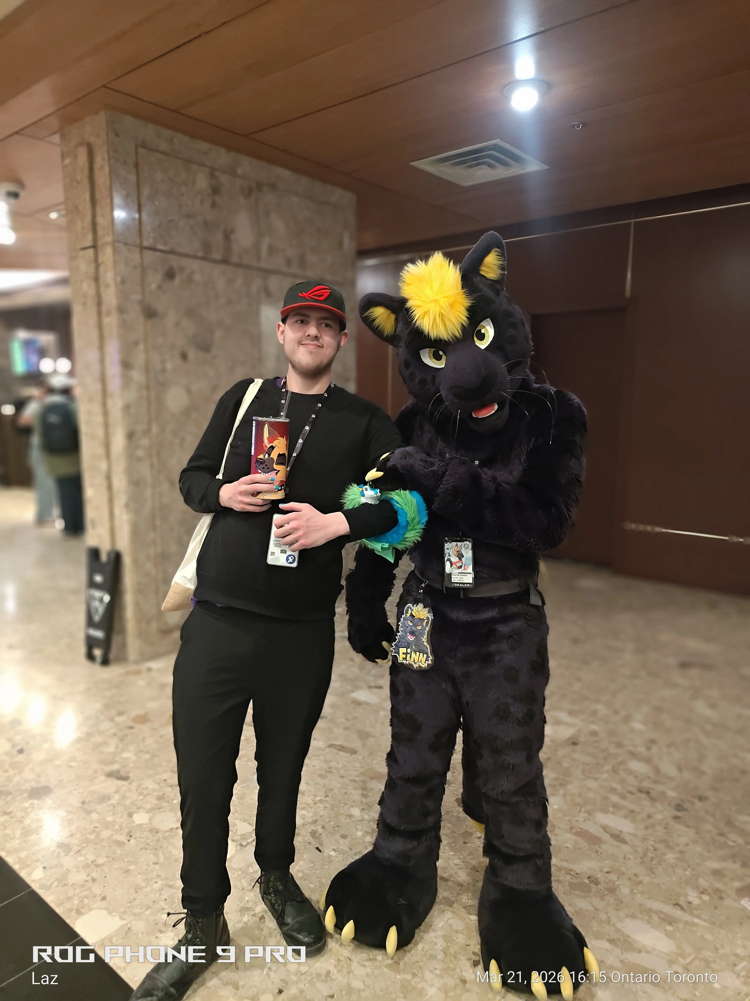 A man in black clothes and a black hat posing with a person in a black panther costume with yellow accents, in an indoor setting with a stone wall and wooden ceiling.