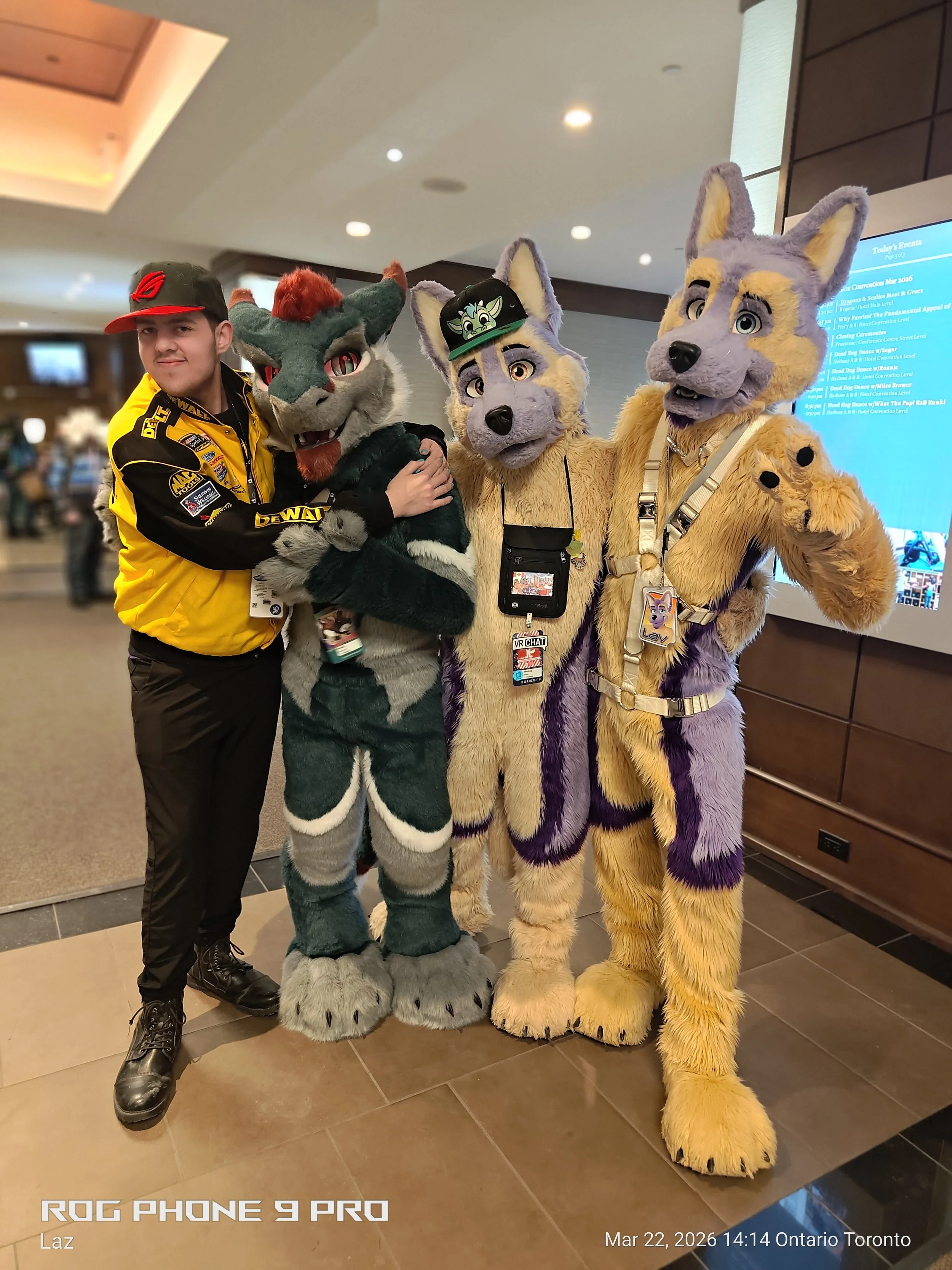 A person in a yellow and black racing jacket and black boots standing next to three people dressed in furry mascot costumes resembling animals, with the background showing a lobby area with a digital event schedule display.