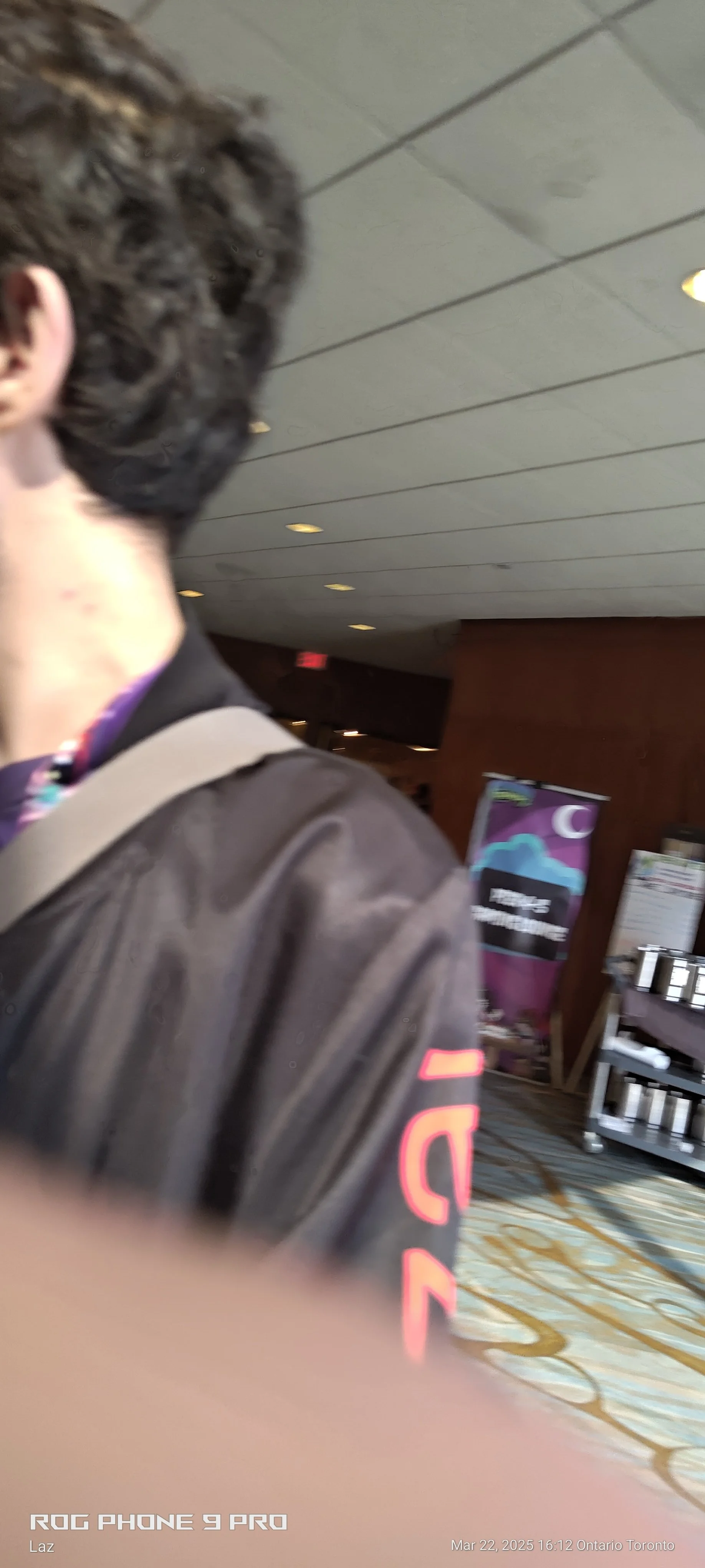 Part of a person's face and shoulder, with curly dark hair, wearing a black jacket and a backpack strap, in an indoor setting with ceiling lights and a banner in the background.
