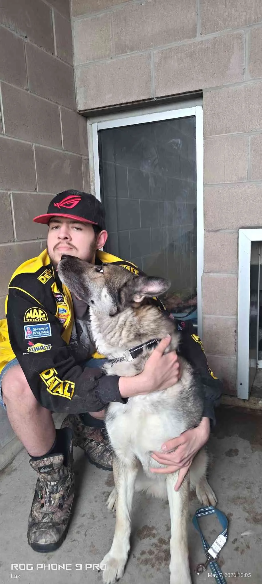A young man in a racing jacket and a cap is sitting against a brick wall while hugging and kissing a large Siberian Husky dog.