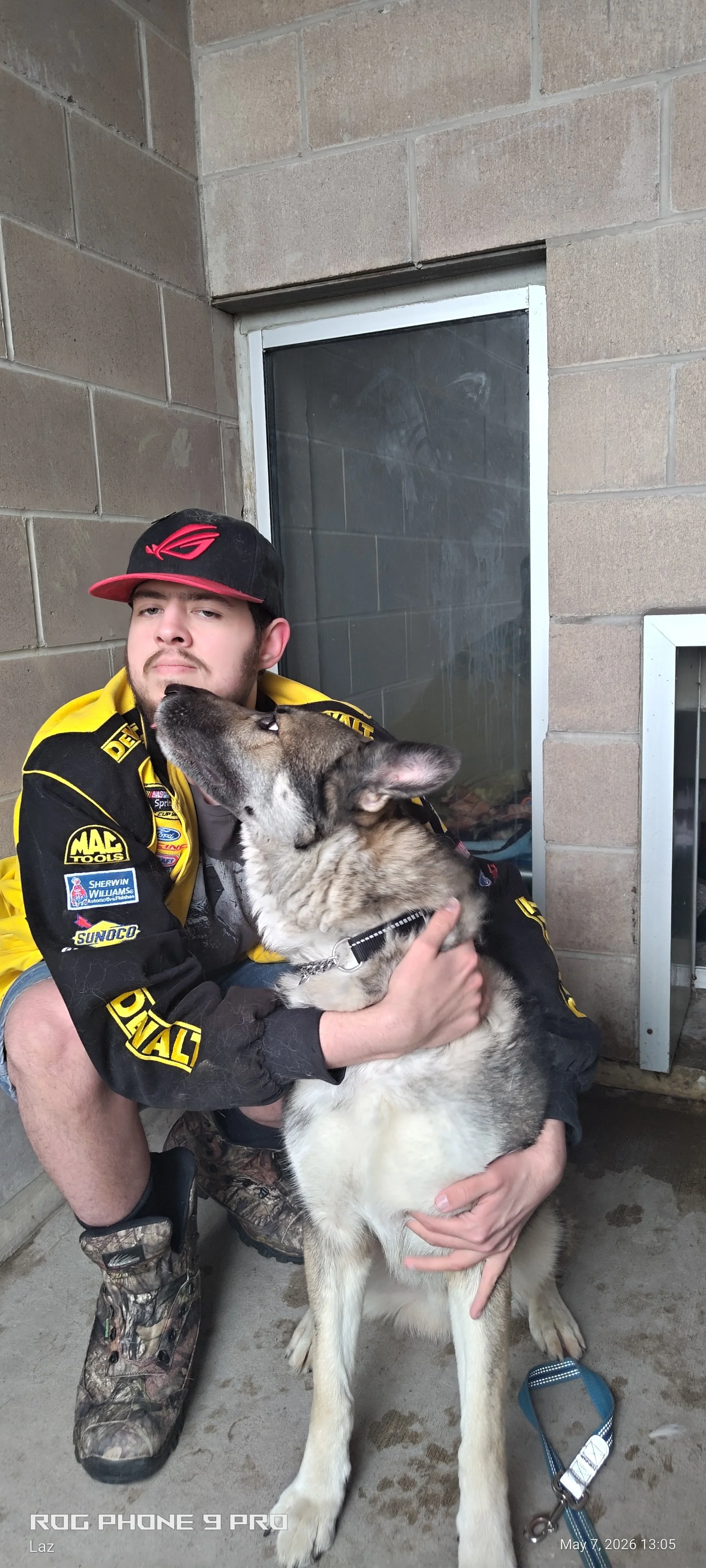 A man in a black and yellow racing jacket and camouflage boots sitting on a concrete floor, hugging a large shepherd dog, with the dog licking his face. The setting appears to be indoors with a brick wall and a small window behind them.