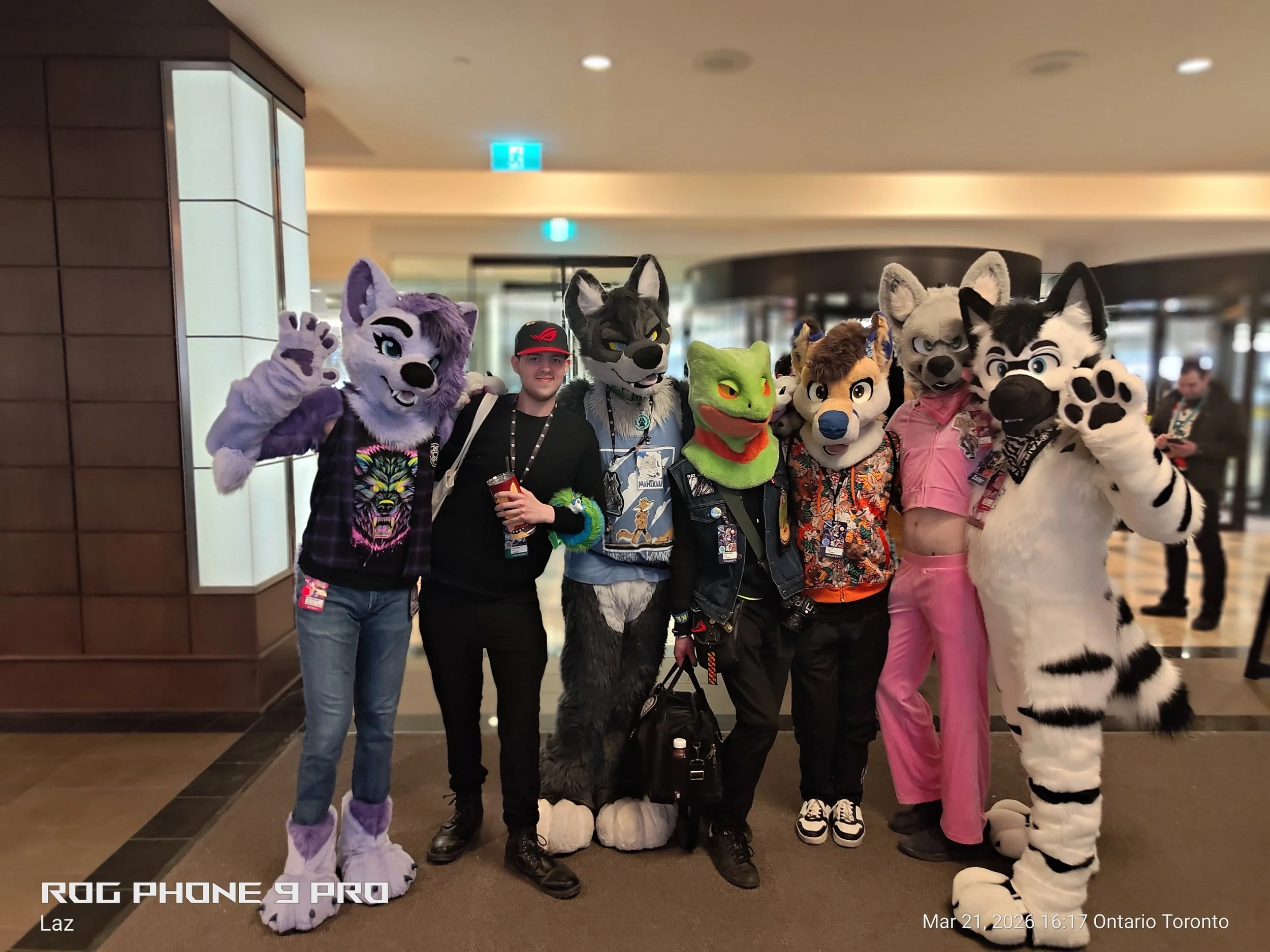 A group of people in fursuits and costumes posing for a photo in a public indoor space. The group includes characters resembling anthropomorphic animals, such as wolves, a frog, and other furry characters, standing close together and smiling.