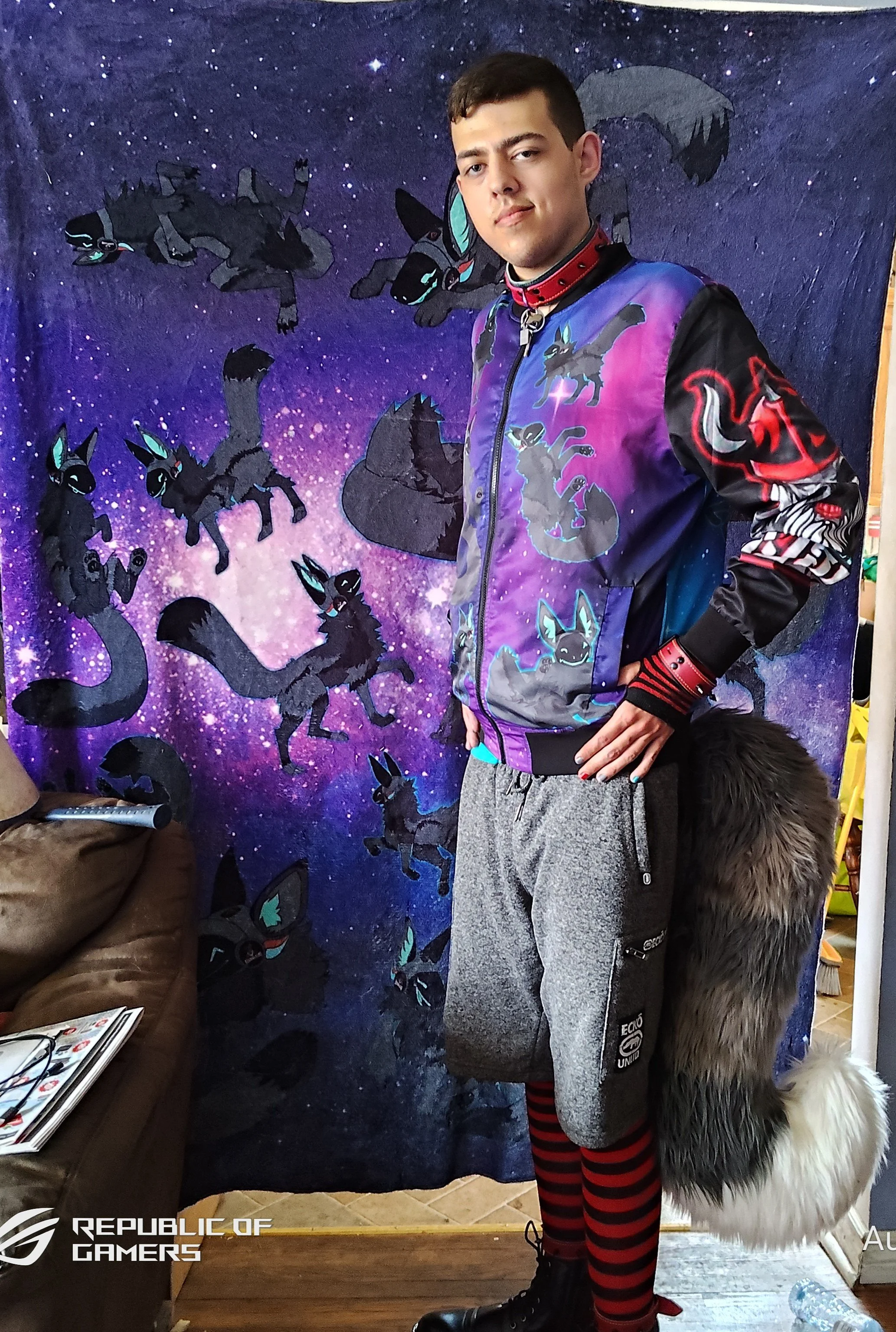 A young man standing indoors in front of a backdrop decorated with characters from Pokémon, such as Umbreon. He is wearing a colorful jacket with Pokémon designs, grey sweatpants, and red and black striped socks. Part of a furry costume is visible on