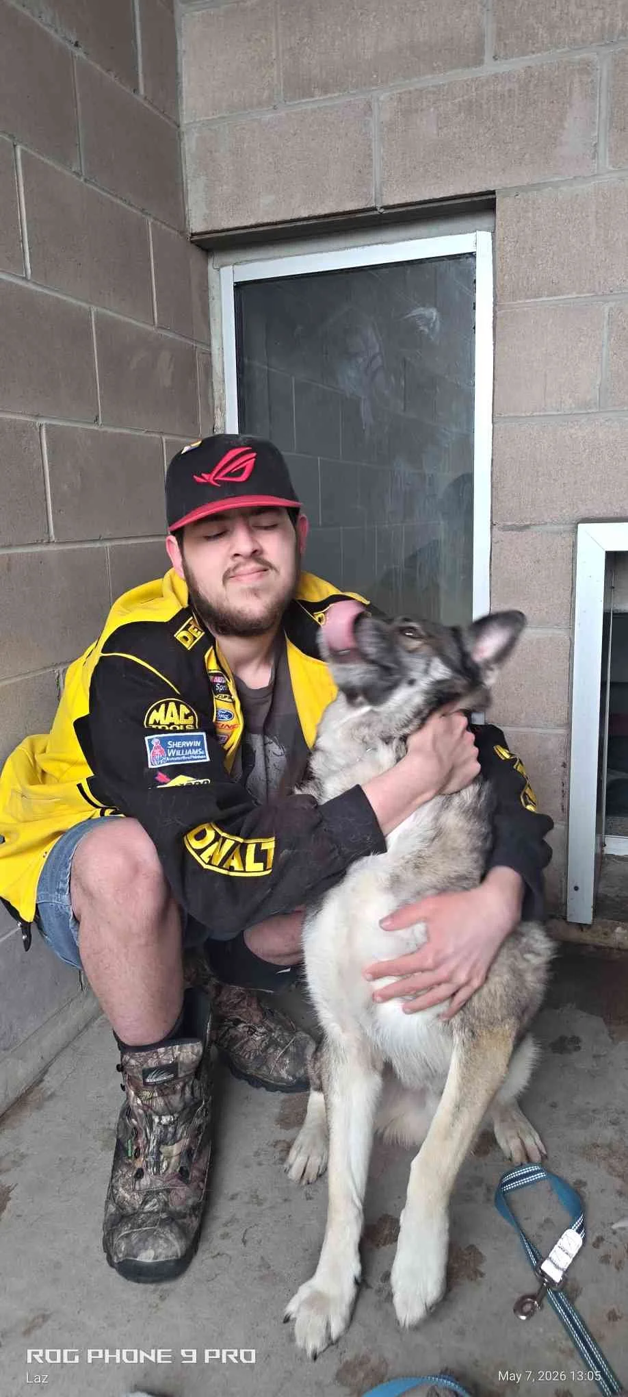 A young man wearing a black and yellow racing jacket, a cap, and camouflage boots is sitting on the ground, hugging a large, fluffy Siberian Husky dog. The dog is looking up and licking the young man's face. They are in an industrial area with brick 