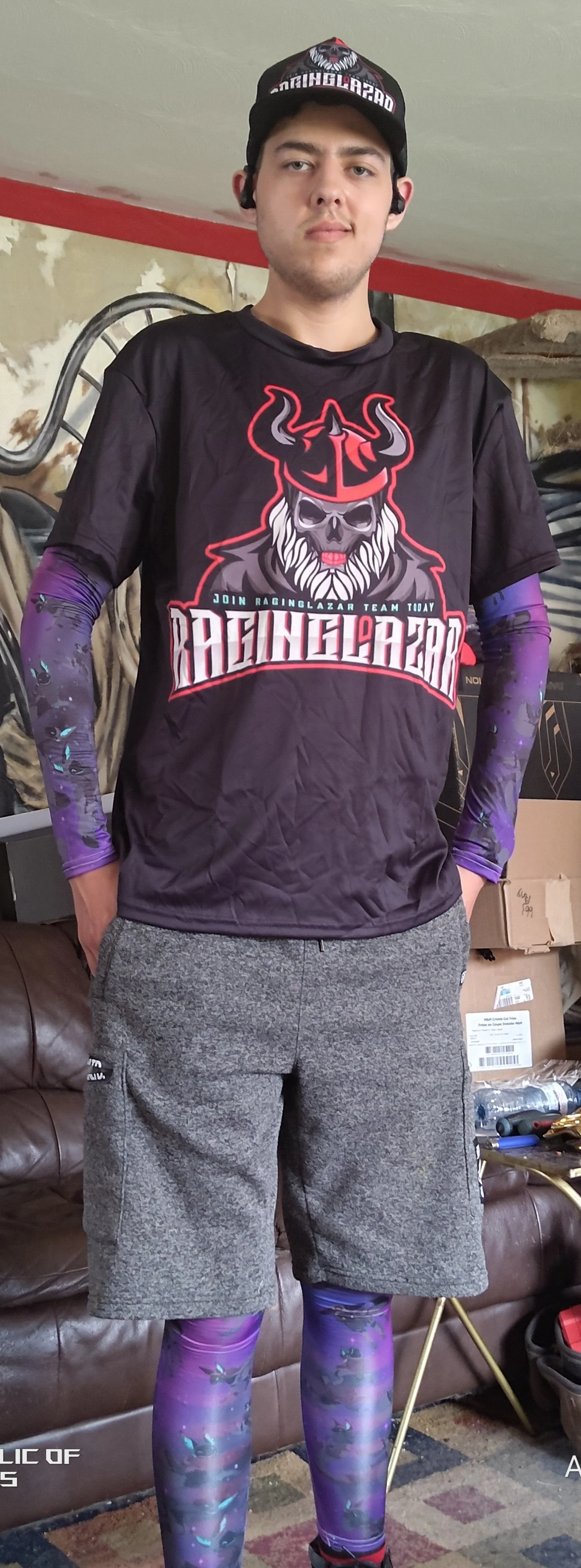 Young man standing indoors, wearing a black and purple sports jersey, gray shorts, purple and black patterned leggings, a black cap with a graphic, and wireless earbuds.