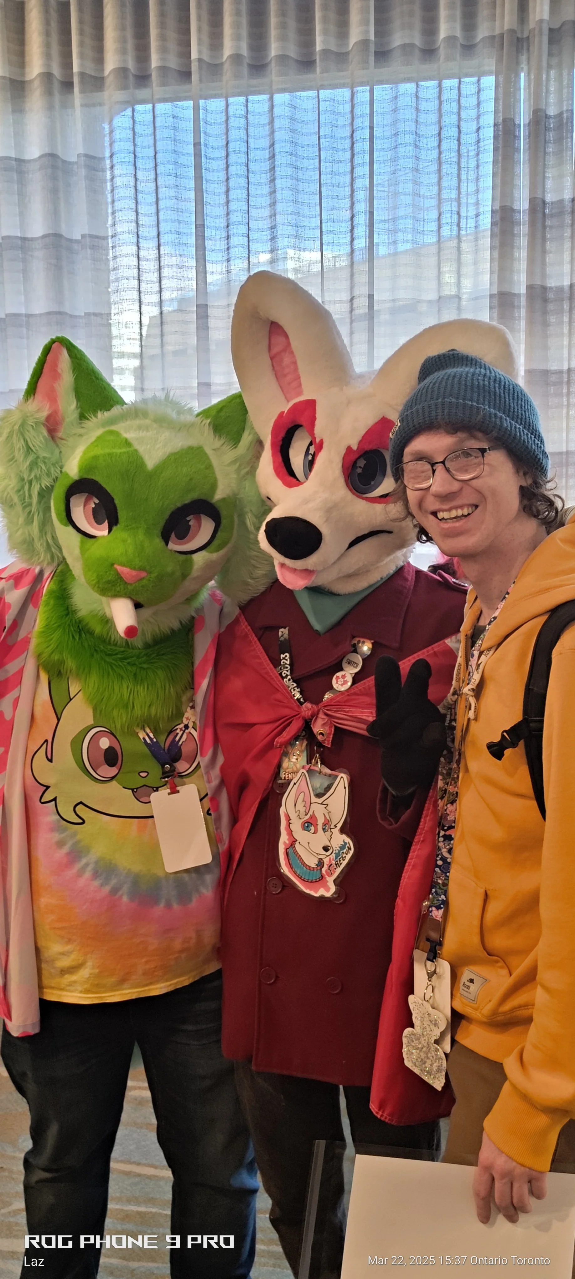 Person dressed in a green furry animal costume with big eyes and a tie-dye shirt, standing next to a person in a large white fursuit with pink accents and a black nose, and another person wearing glasses, a blue beanie, and a yellow jacket, all smili