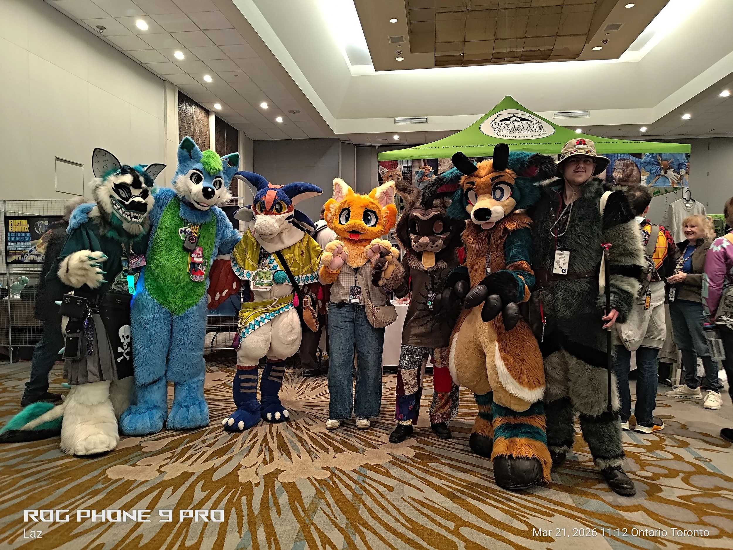 Group of people dressed in colorful furry animal costumes, representing various species, standing in a line at an indoor event. Behind them is a green canopy banner with the logo of the Toronto Wilderness Committee. The setting appears to be a conven