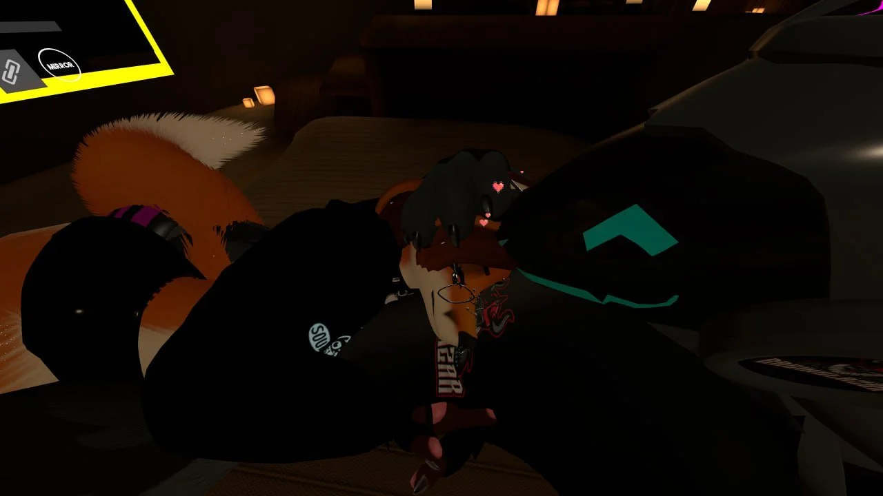 A person with dark hair, glasses, and wearing a black jacket with patches, lying on a bed with their head resting on another person’s arm, in a dimly lit room with a computer screen visible in the background.