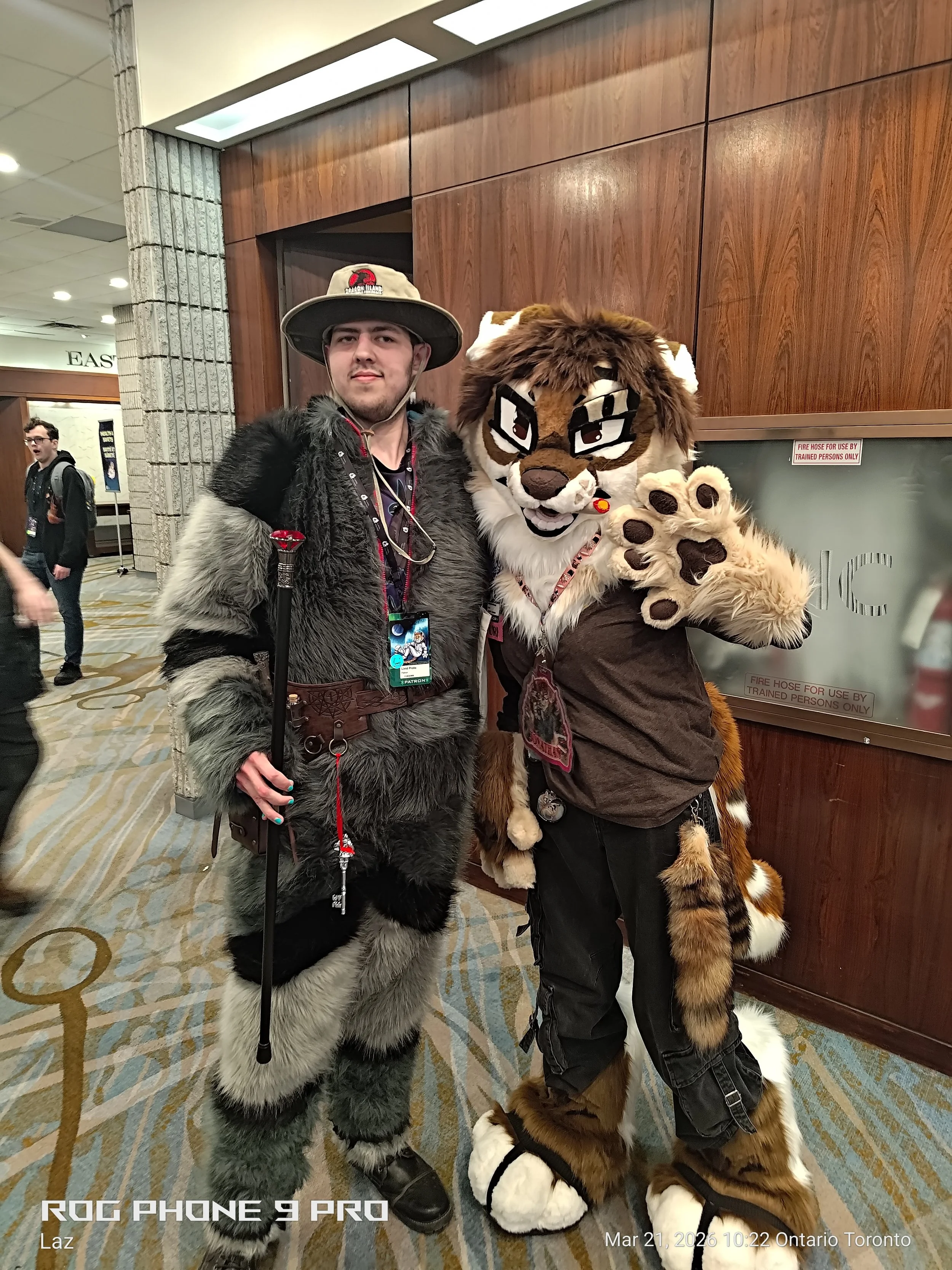 Two individuals dressed in fur and animal-themed costumes are posing together indoors. One person is dressed as a wolf or dog with gray and black fur and a wide-brimmed hat. The other is dressed as a lion or tiger with brown, tan, and white fur, glas