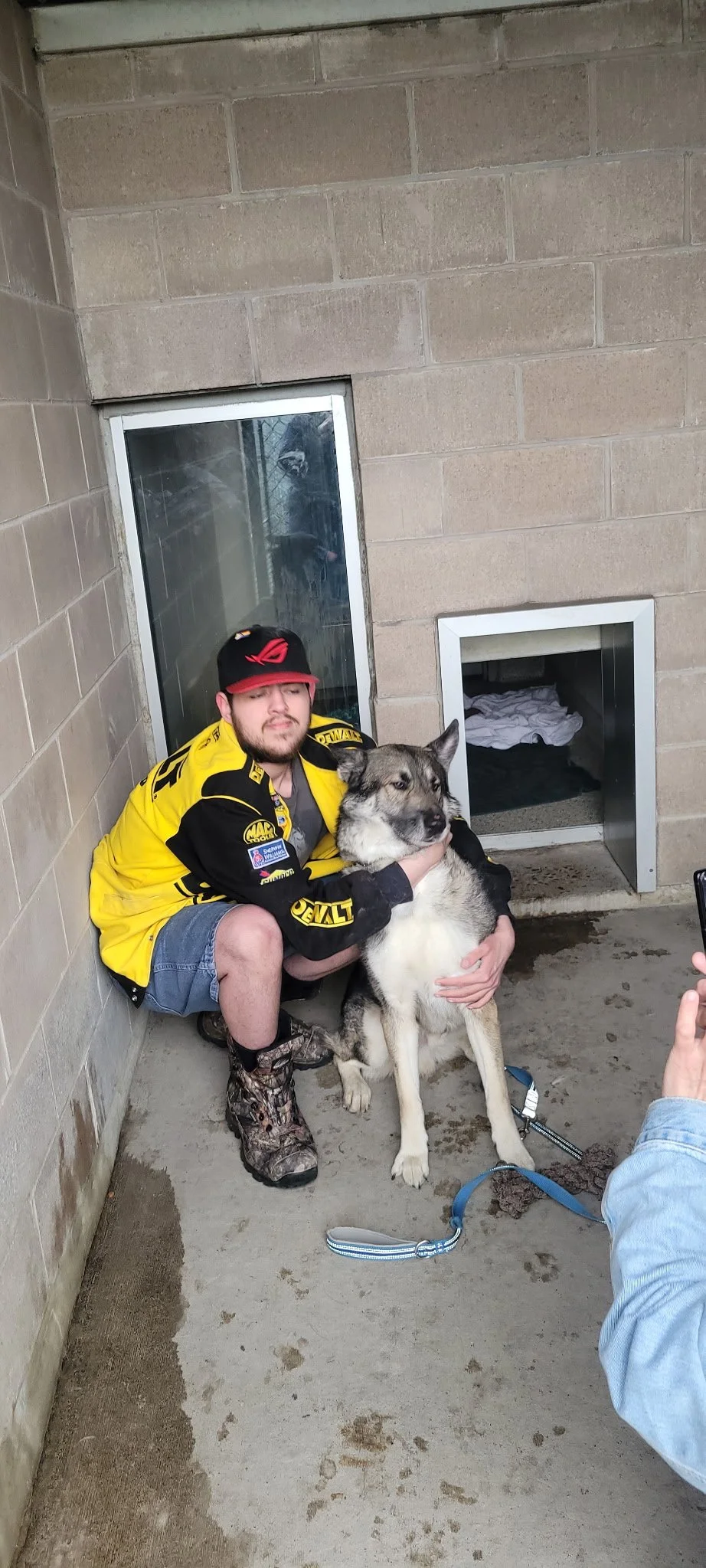 A man wearing a yellow and black racing jacket and shorts, squatting and holding a large dog, a Siberian Husky, inside a concrete shelter. There is a window and a small pet door in the wall behind him. The ground appears damp with some patches of wat