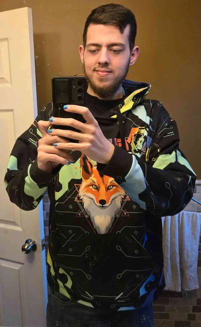 A young man with short brown hair and a beard taking a mirror selfie with a black smartphone. He is wearing a black hoodie with neon green and blue accents, featuring a graphic fox design on the front.