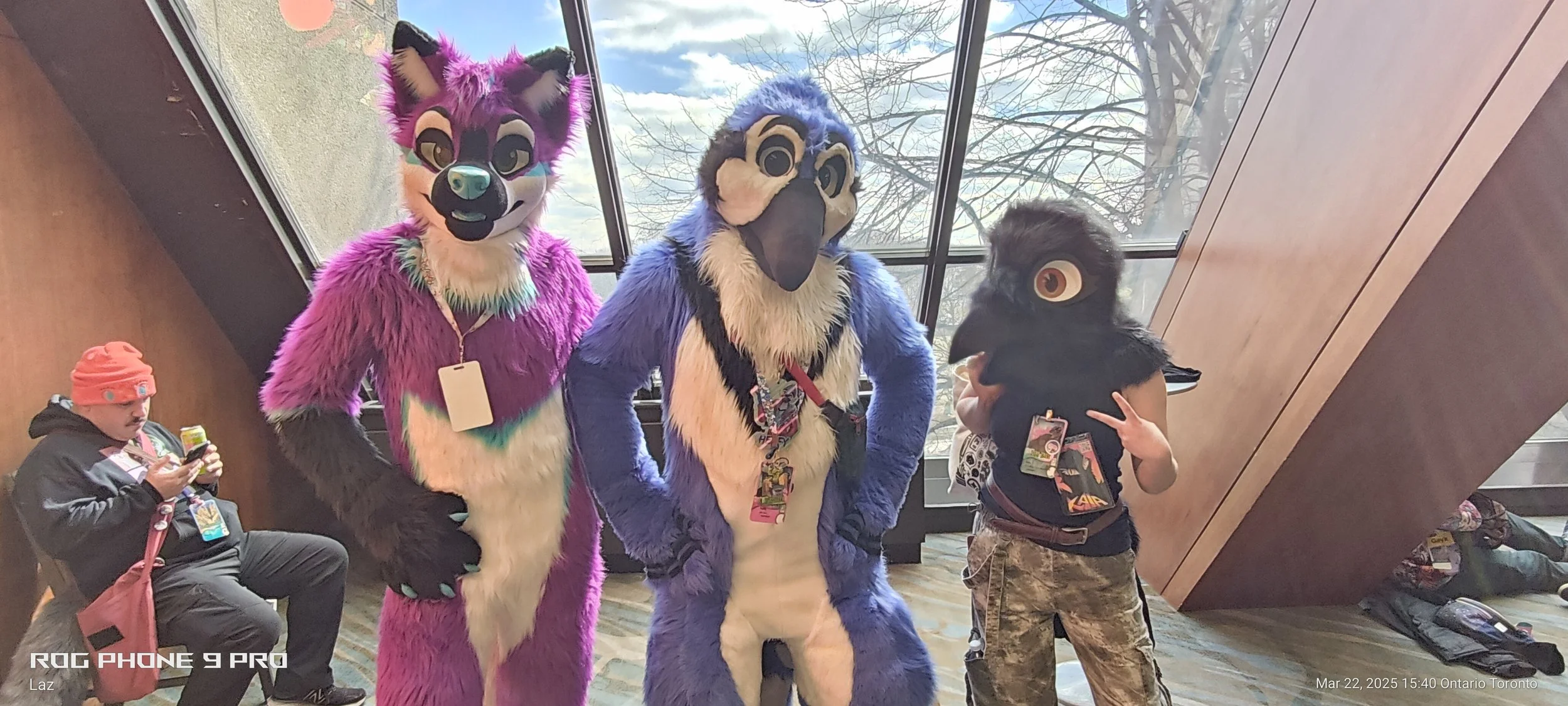 Three people dressed in colorful furry animal costumes, with two children nearby. The costumes include a purple fox, a blue and white bird, and a black bird. They are indoors near large windows with a view of trees outside.