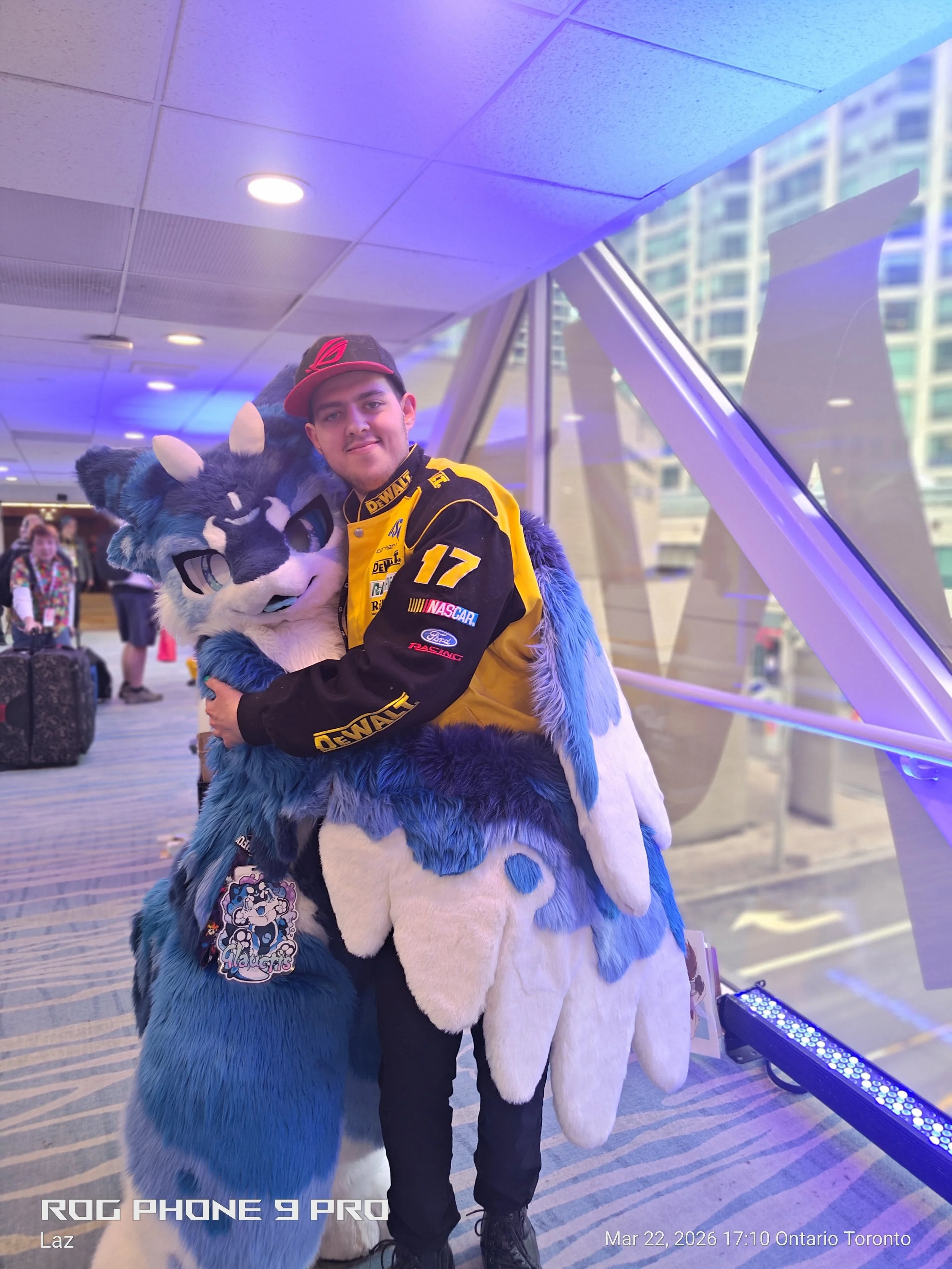 A young man smiling and hugging a person in a blue and white furry costume with horns inside a modern building with large windows and high-rise buildings outside.