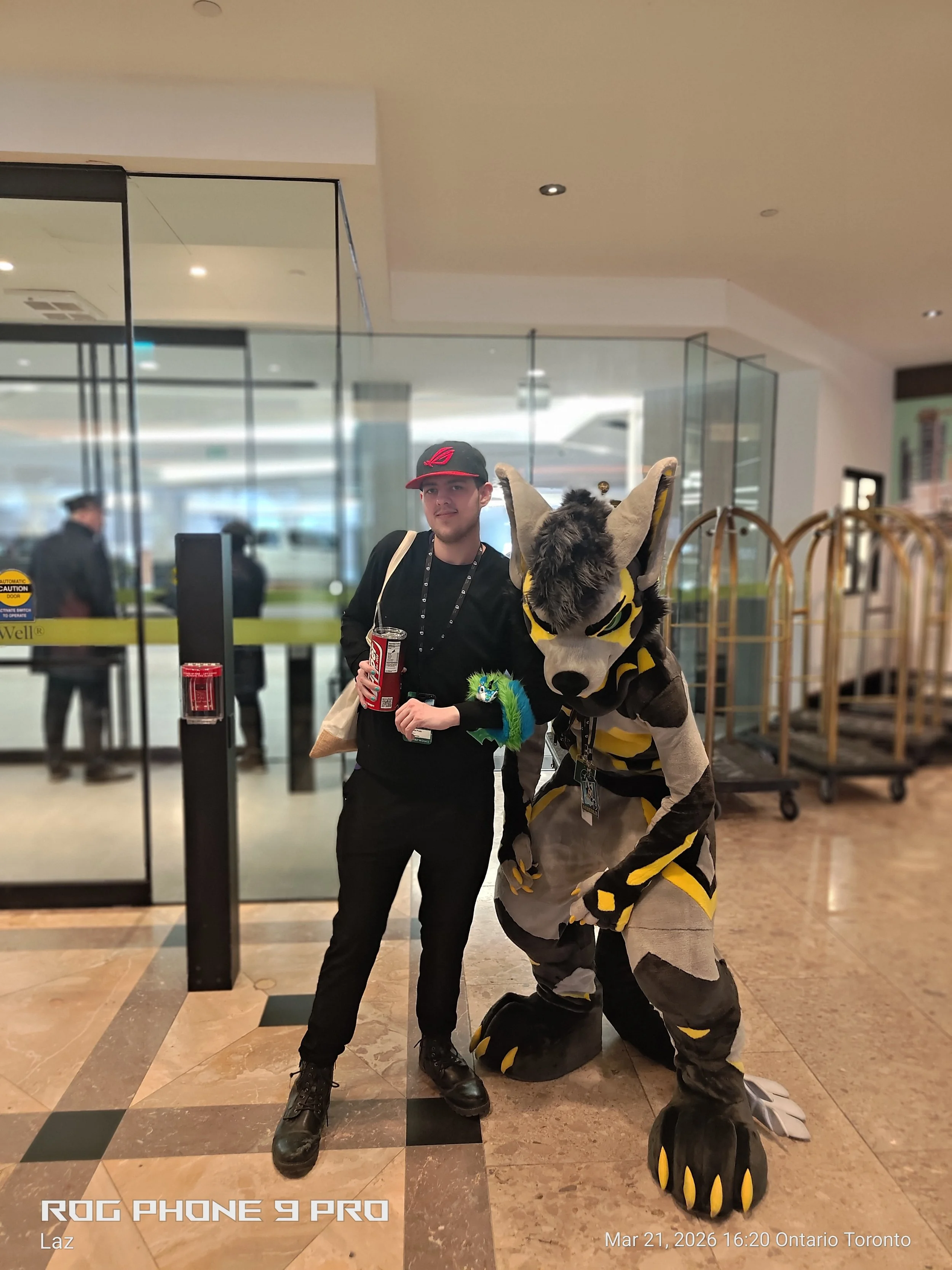 A man in black clothing and a red and black cap standing next to a person in a tiger costume inside a building near a glass door.