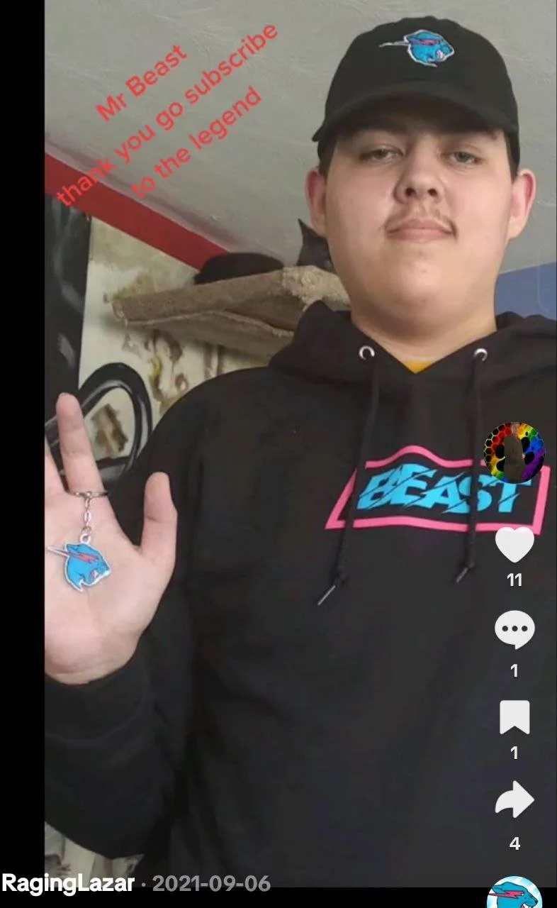 A young man wearing a black cap with a blue car logo, a black hoodie with a pink and blue 'Beast' logo, and a keychain with a blue creature emblem hanging from his hand. He is making a peace sign with his right hand. There is red text in the upper le