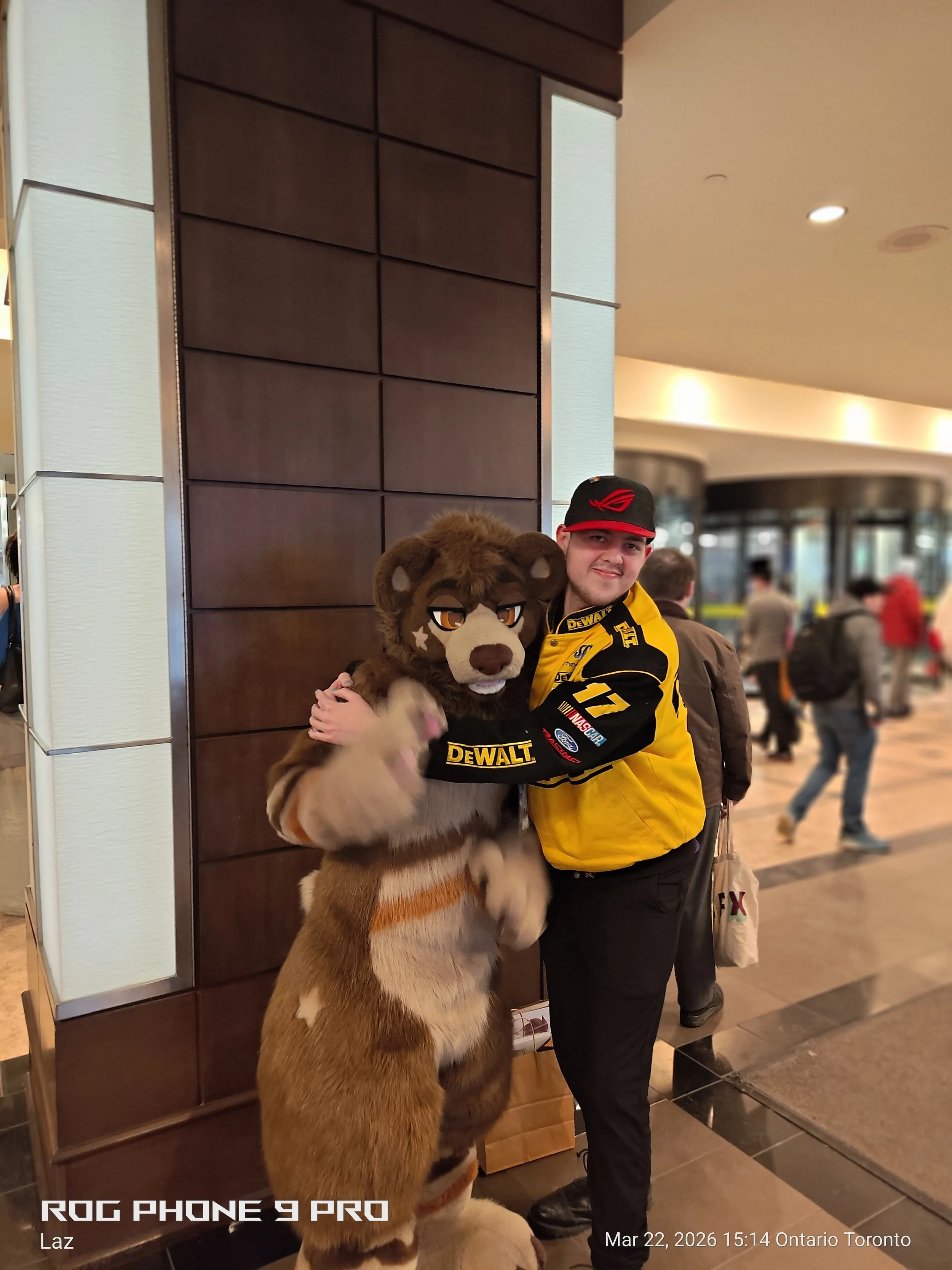 A person in a yellow racing jacket and a red and black hat hugging a person in a lion mascot costume in a public indoor space.