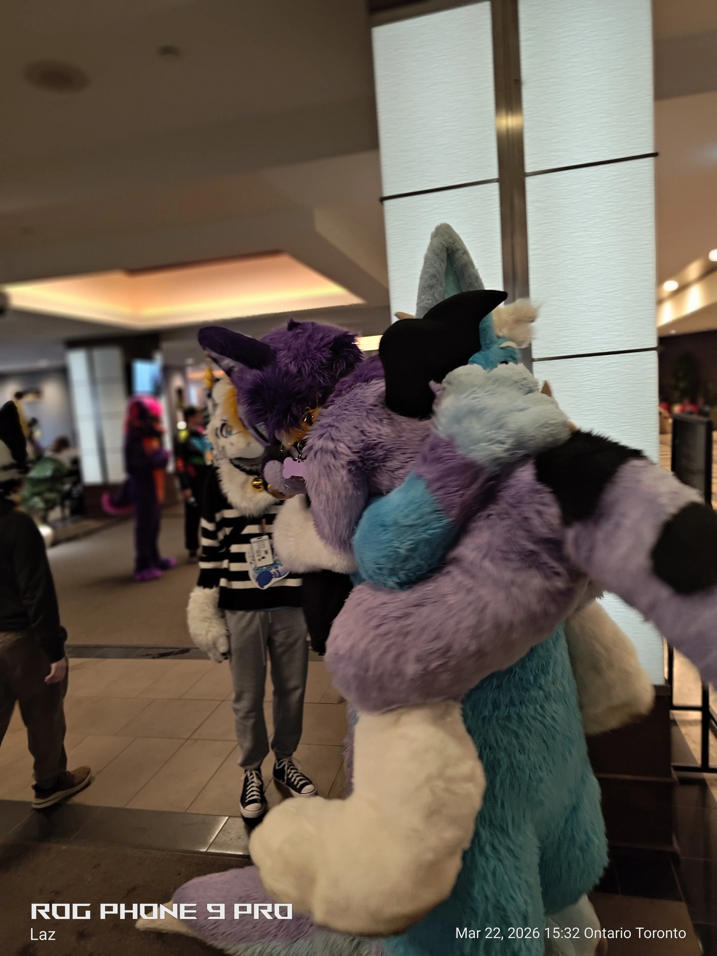 A person dressed in a colorful, furry animal costume with purple, blue, and white fur, and wearing black wings, is interacting with people in a busy indoor environment, likely at a convention or event.