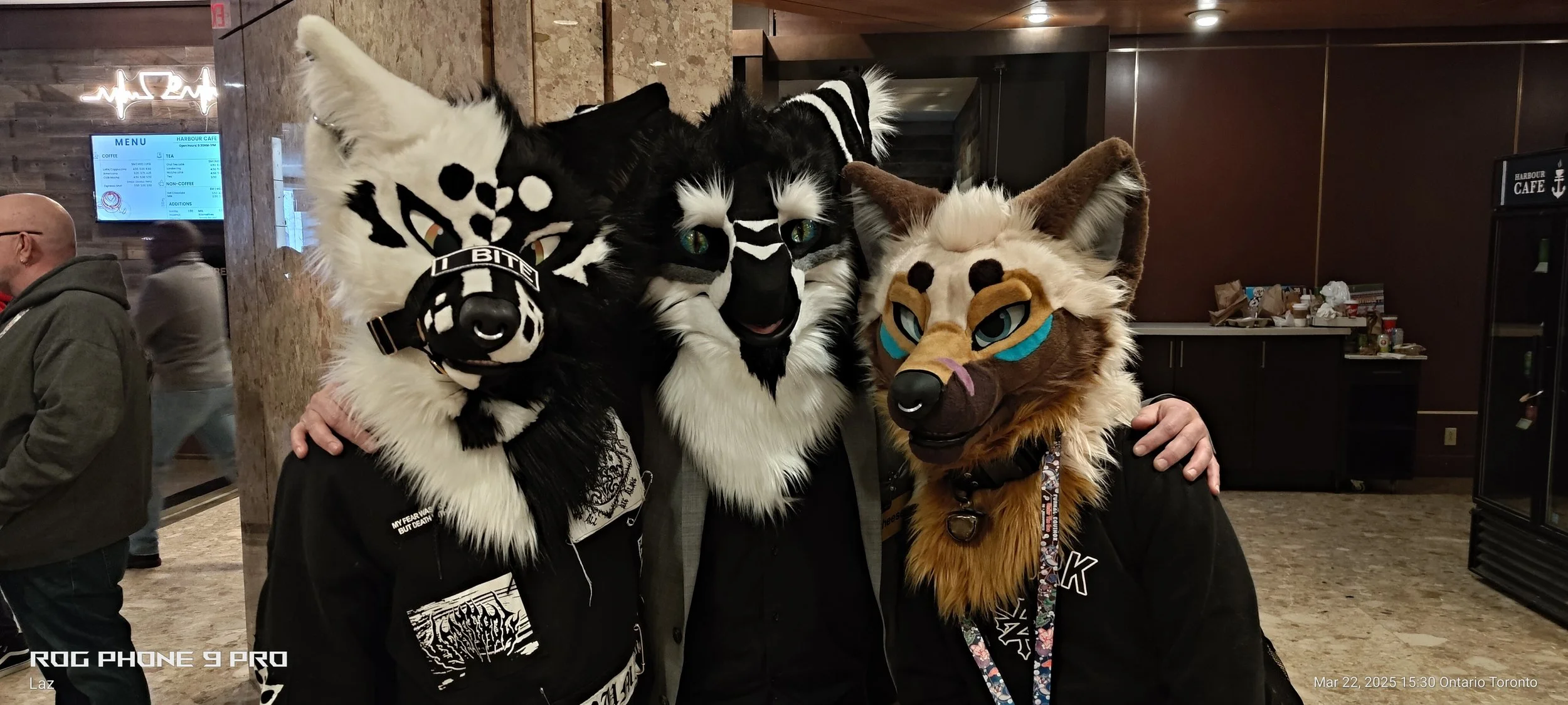 Three people wearing detailed animal costumes and masks, resembling a skunk, a wolf, and a fox, standing close together in an indoor setting with other people and a menu in the background.