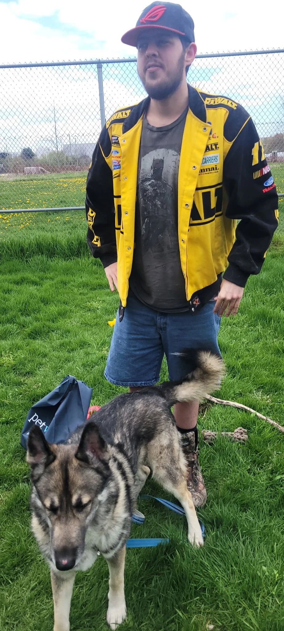 A man wearing a black and yellow racing jacket, a cap, and shorts standing outside on grass with a husky dog on a leash in front of him.