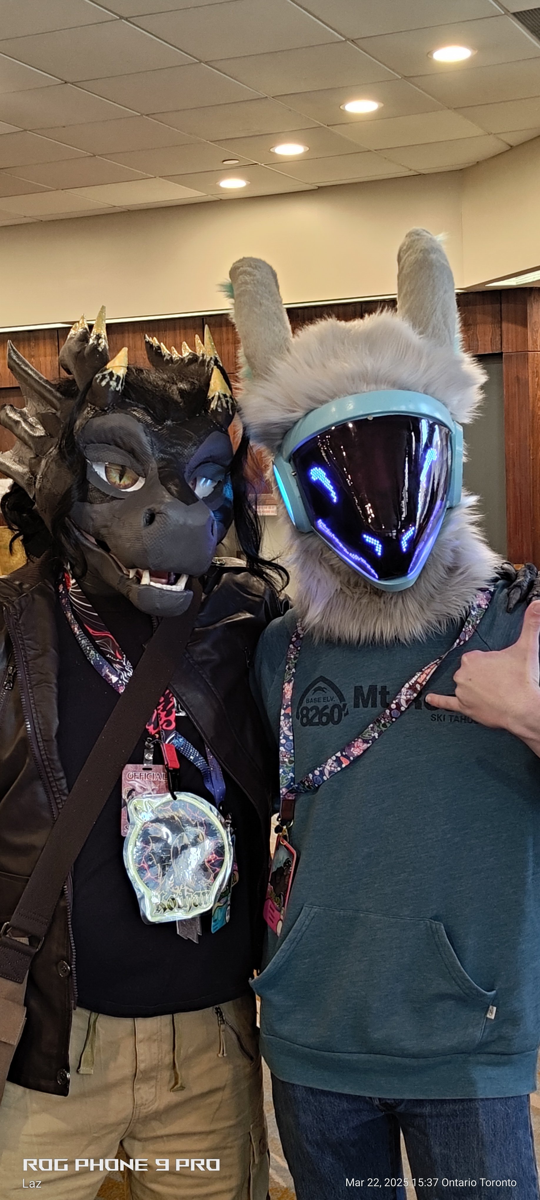 Two people wearing elaborate masks: one with a dragon mask and another with a futuristic helmet, taking a selfie indoors.