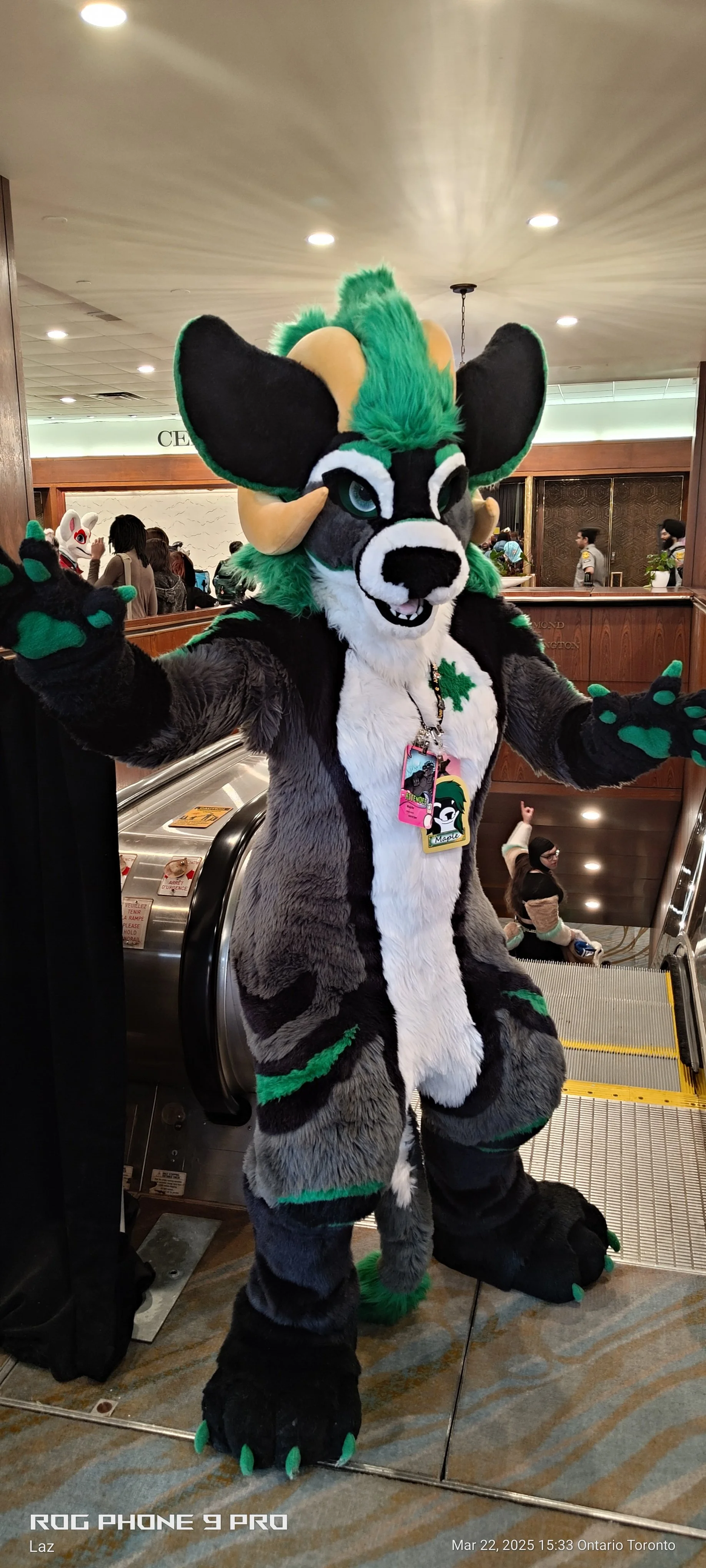 A person dressed in a colorful fursuit of a cartoon fox character, standing at an event indoors, with a crowd in the background and an escalator nearby.