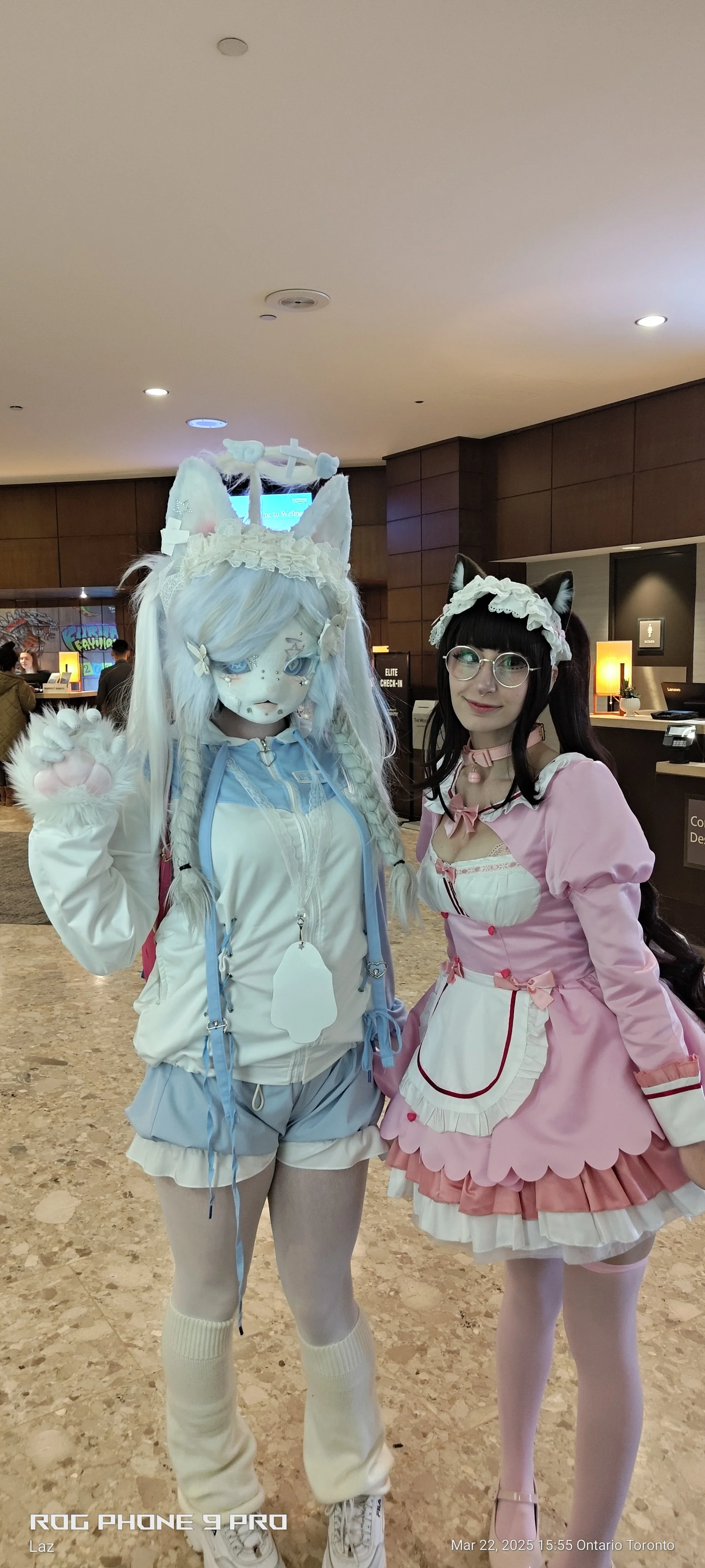 Two women dressed in elaborate costumes pose in a lobby. One is dressed as a blue and white fantasy character with animal ears and paws, the other as a pink maid with cat ears, glasses, and a lace headband.