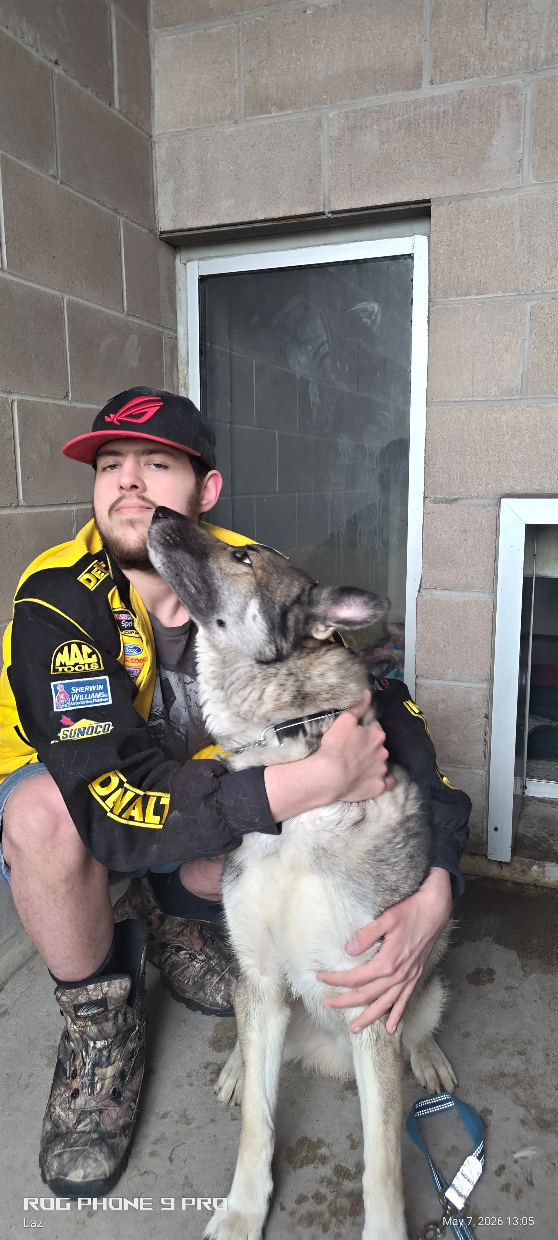 A man wearing a black and red cap and a yellow racing jacket sitting on the ground hugging a large husky dog.