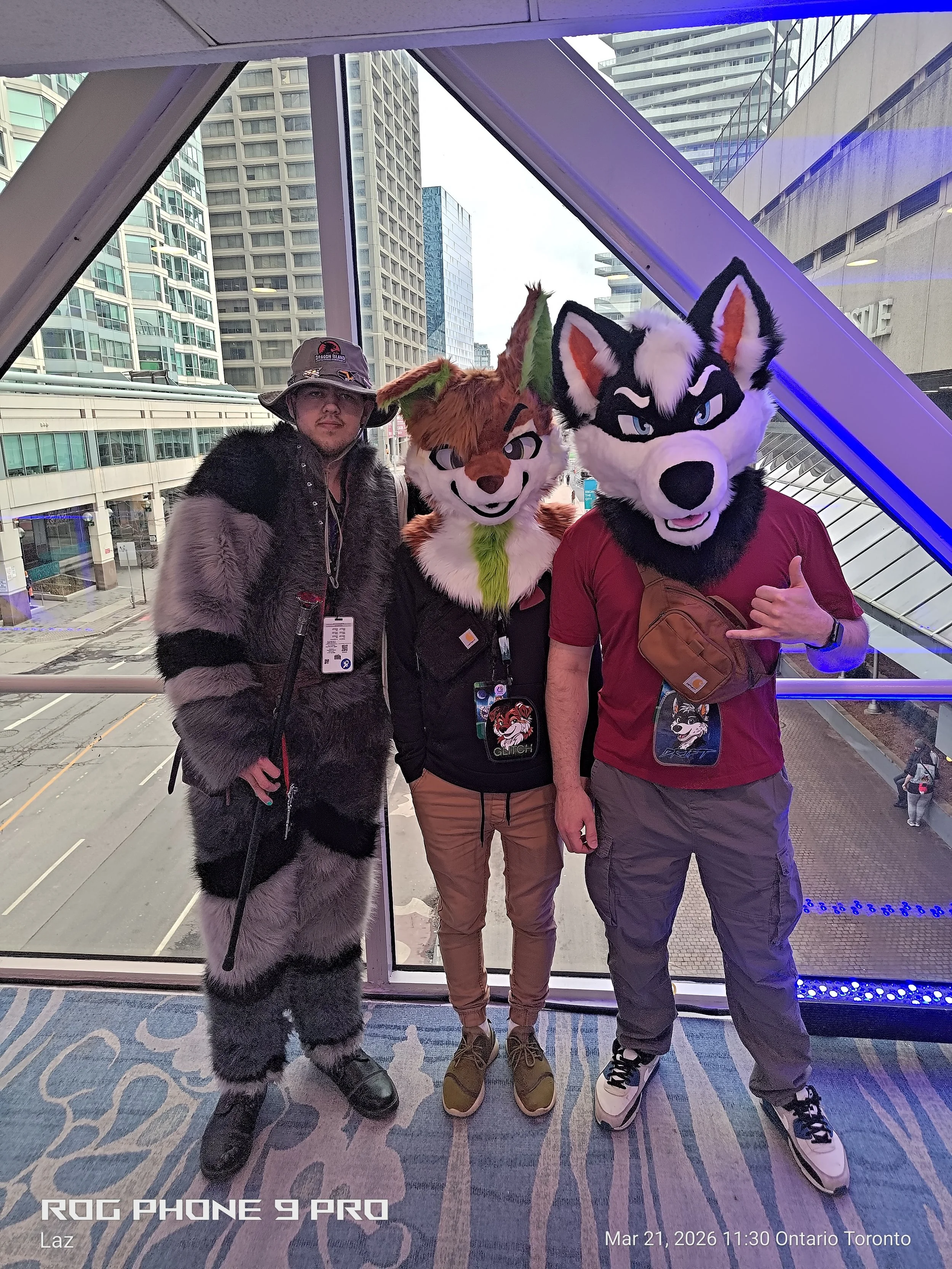 Three individuals dressed in furry costumes posing together in an urban setting with tall buildings in the background. The person on the left is in a cougar costume, the middle person in a fox costume, and the person on the right in a wolf costume.
