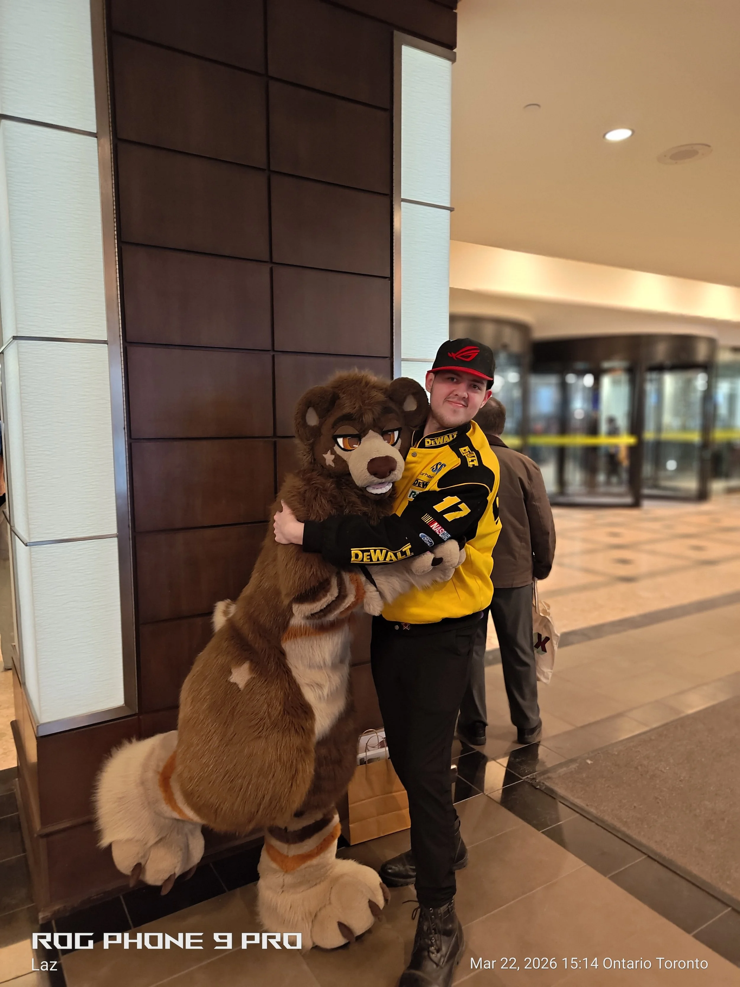A young man dressed in a yellow racing jersey with black and red accents, standing inside a building and hugging a person in a lion mascot costume. The mascot has a detailed face and a large mane. Thebackground includes glass doors and a tiled floor,