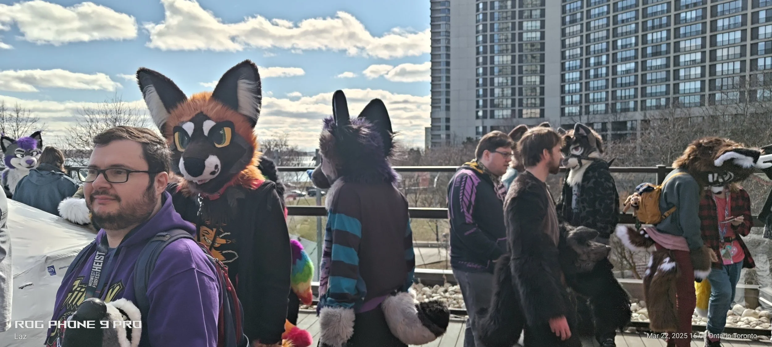 People dressed in furry animal costumes walking outdoors in an urban park with high-rise buildings in the background on a sunny day.
