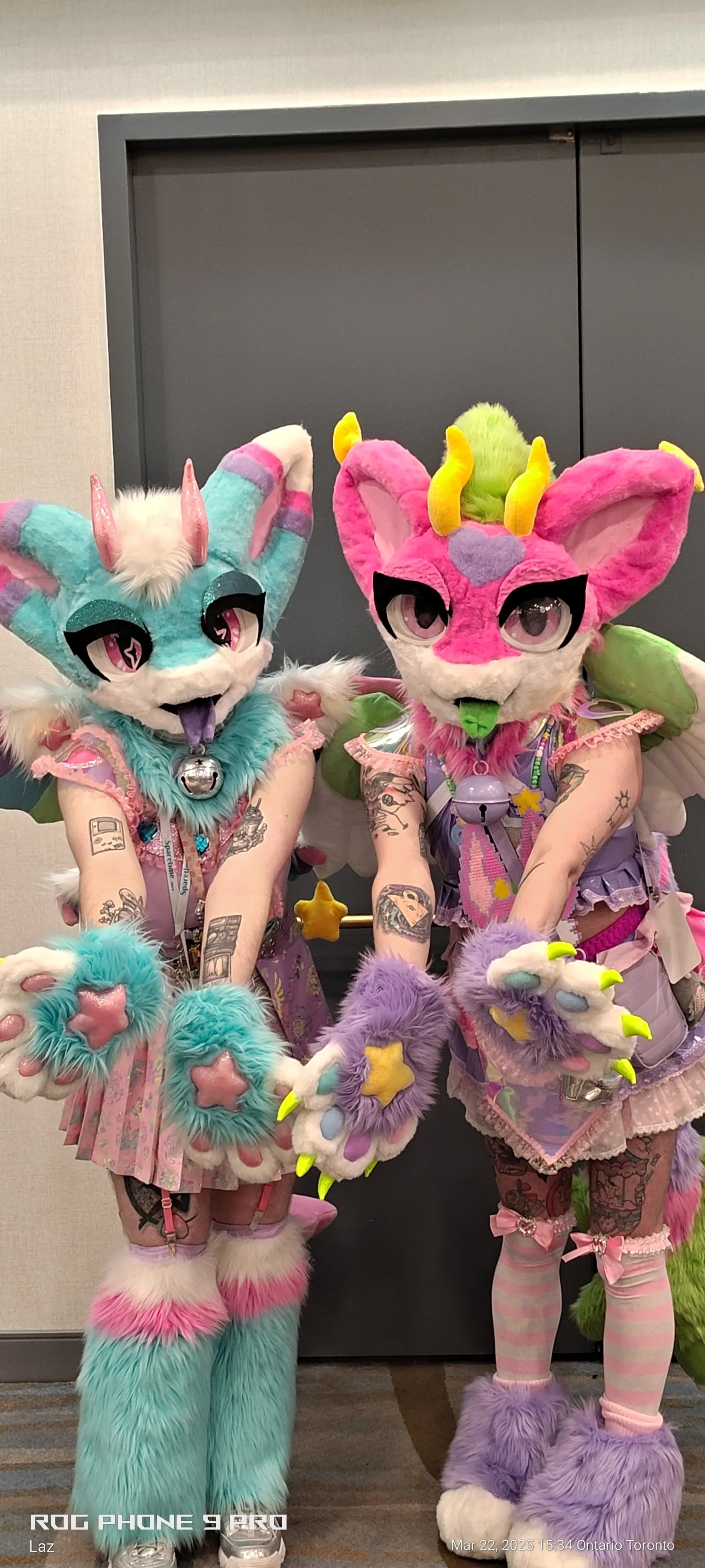 Two people dressed in colorful, furry, pony-themed costumes with large eyes, horns, and wings, standing indoors in front of a gray door.
