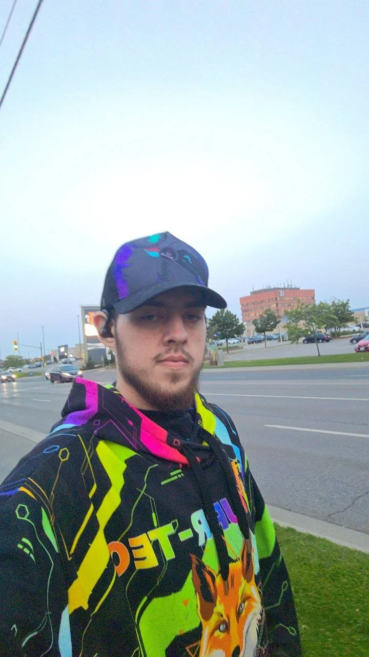 A young man with a beard standing outdoors on a sidewalk near a busy road, wearing a black baseball cap and a colorful hoodie with a fox graphic.